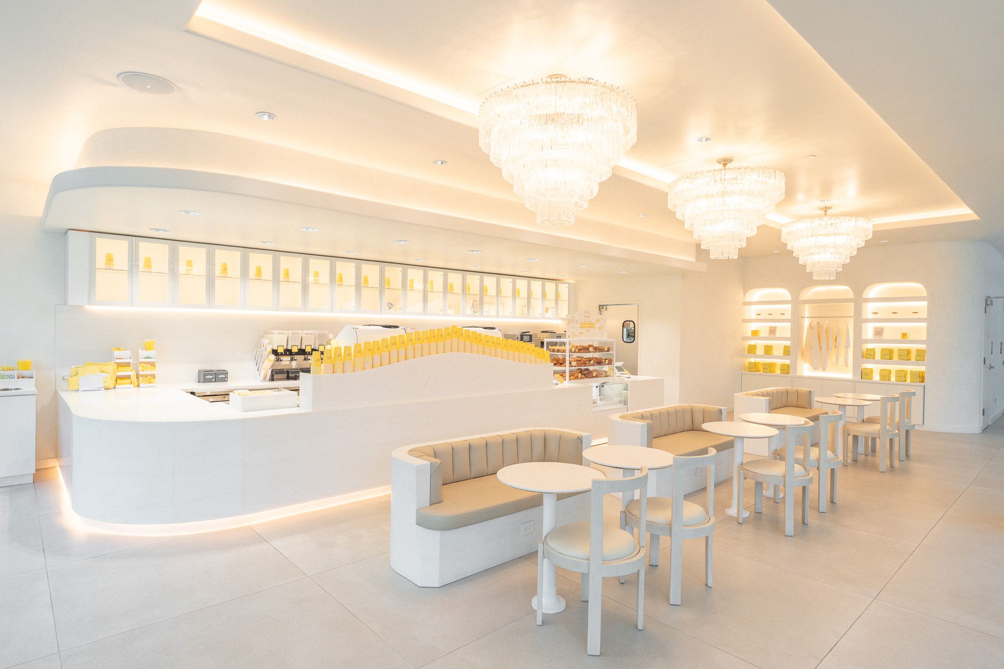 Bright all-white cafe with curved white counter, beige cushioned benches, and small round tables. Yellow product boxes line the counter; large crystal chandeliers hang from a softly lit ceiling.