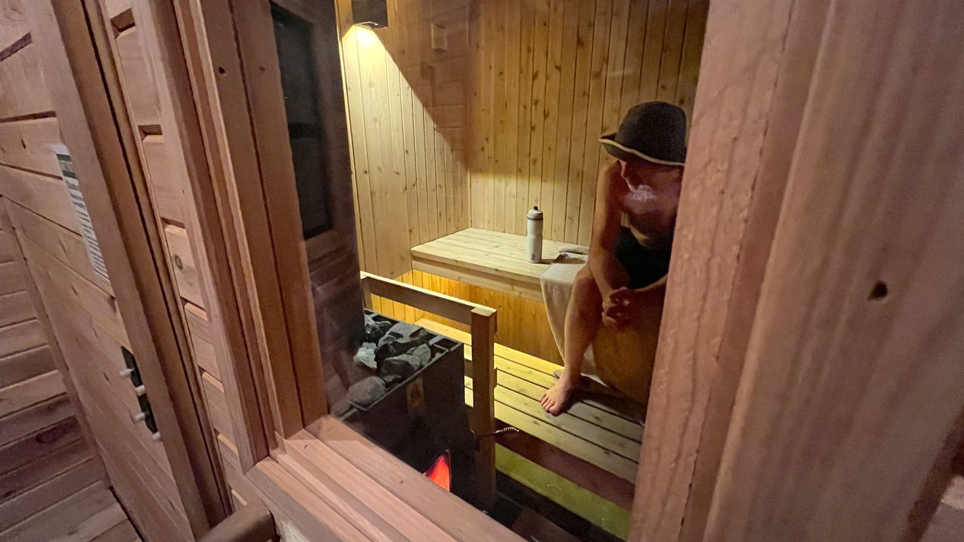 A man photographed through the window of a sauna door. He's wearing a bucket hat and bathing suit and has his hands clasped.