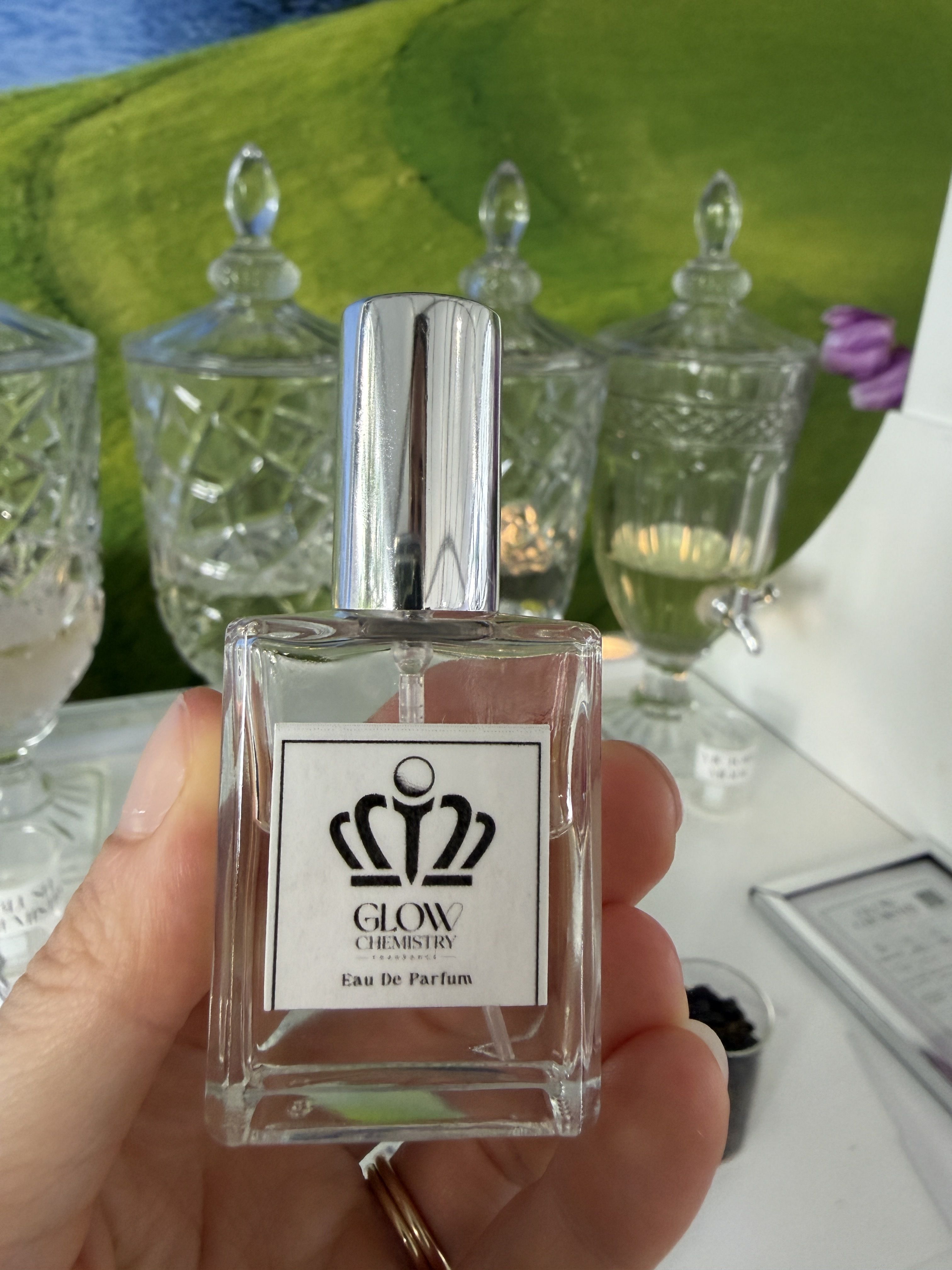 Hand holding a square glass perfume bottle with a tall silver cap; label reads Glow Chemistry and Eau De Parfum, pink liquid inside; blurred crystal jars and a green backdrop behind.