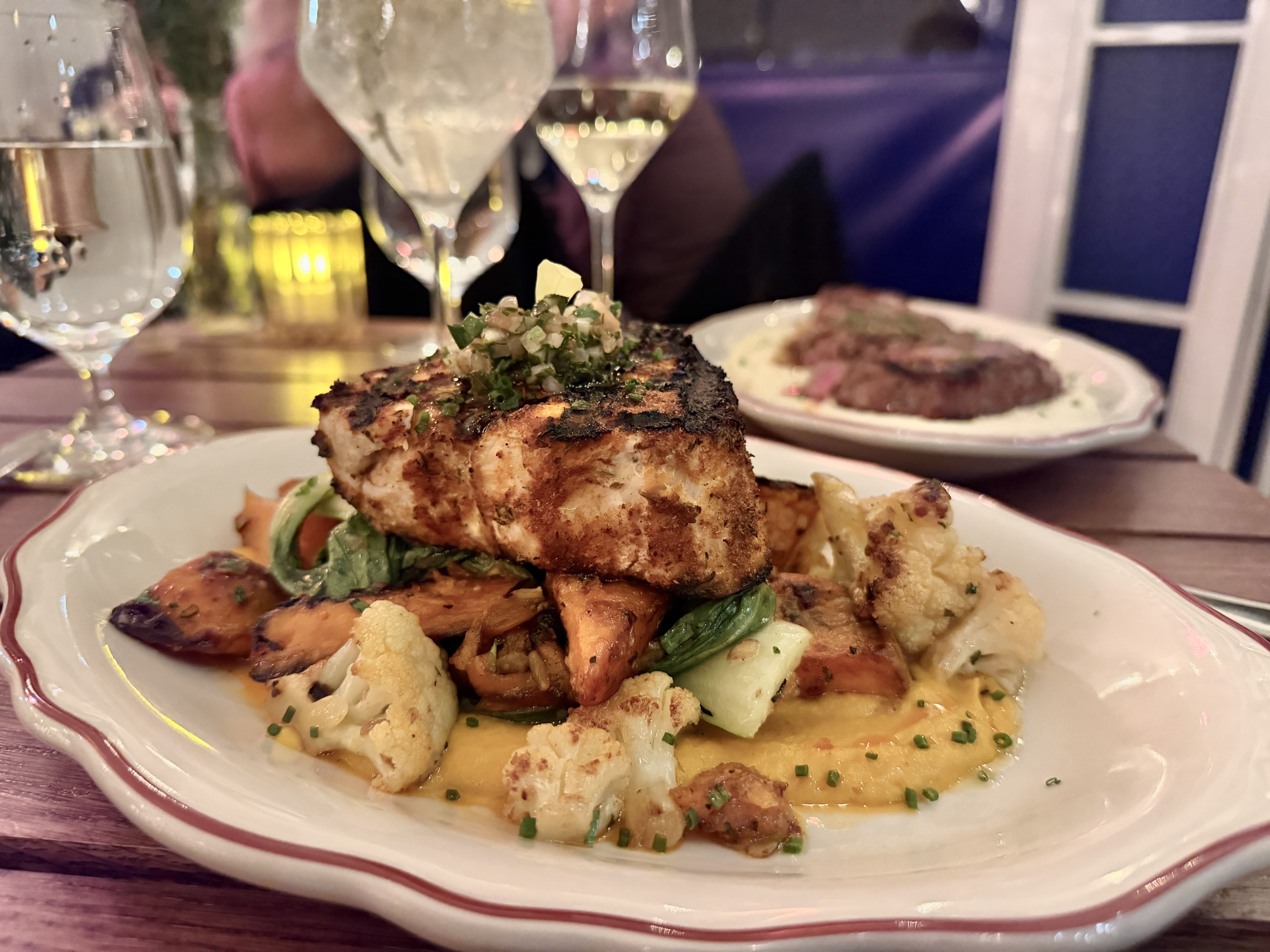 Grilled fish fillet topped with herb sauce, served on roasted vegetables and cauliflower on a white plate with a red rim; wine and water glasses in the background.