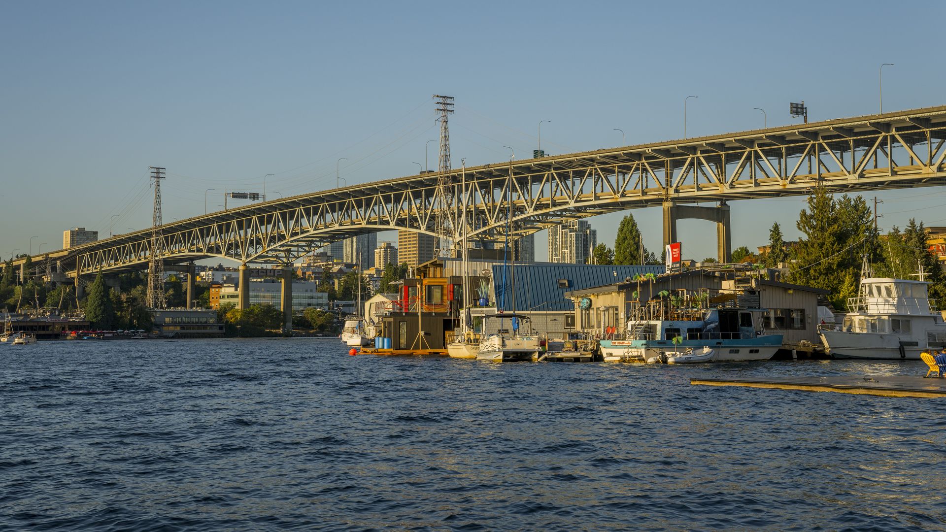 An elevated bridge over water with the Seattle skyline visible in the background and houseboats and floating homes in the foreground.