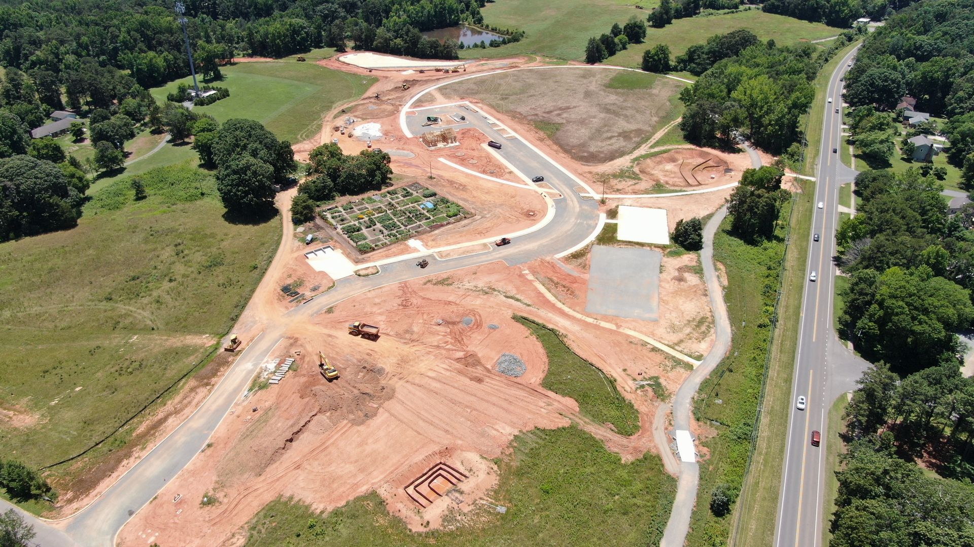 Aerial view of a construction site with red soil, paved roads, vehicles, a garden patch, and surrounding green fields and trees next to a highway with cars.