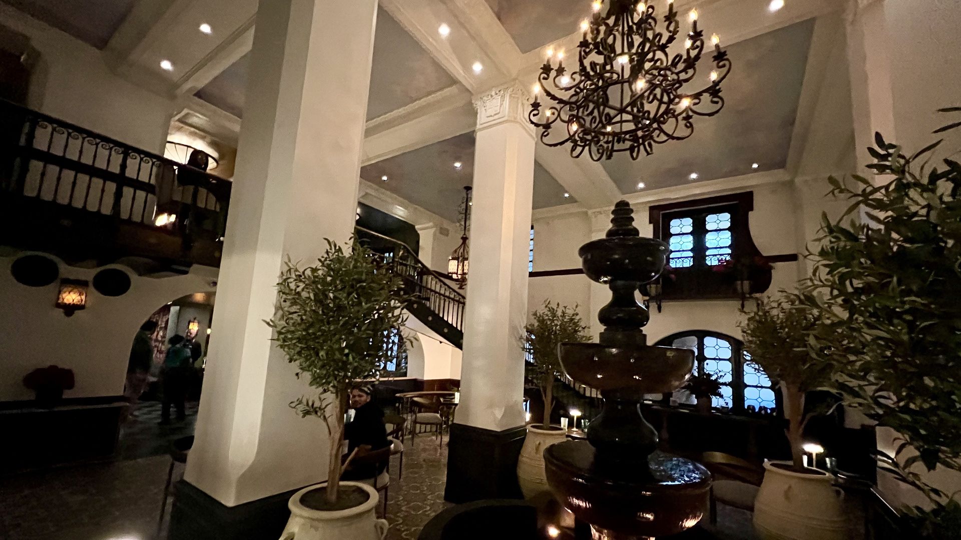 Dimly lit interior of a classic building with white columns, a dark fountain, ornate chandelier, potted plants, arched windows with blue glass, a staircase, and people seated.