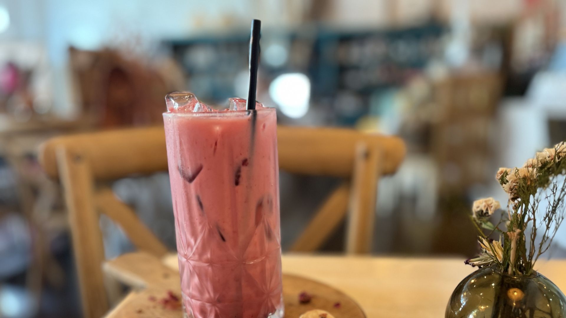 Pink iced drink in a tall glass with ice and a black straw sits on a wooden round cake board; scattered pink petals surround it, with a blurred cafe interior and a dark glass vase of dried flowers nearby.