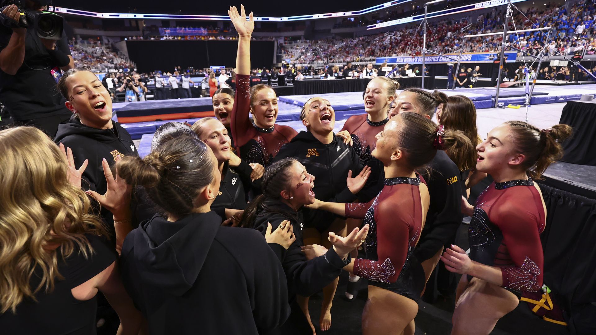 Group of female gymnasts in burgundy and black leotards celebrate in a huddle on the competition floor, as a camera operator films and a crowded arena looks on.
