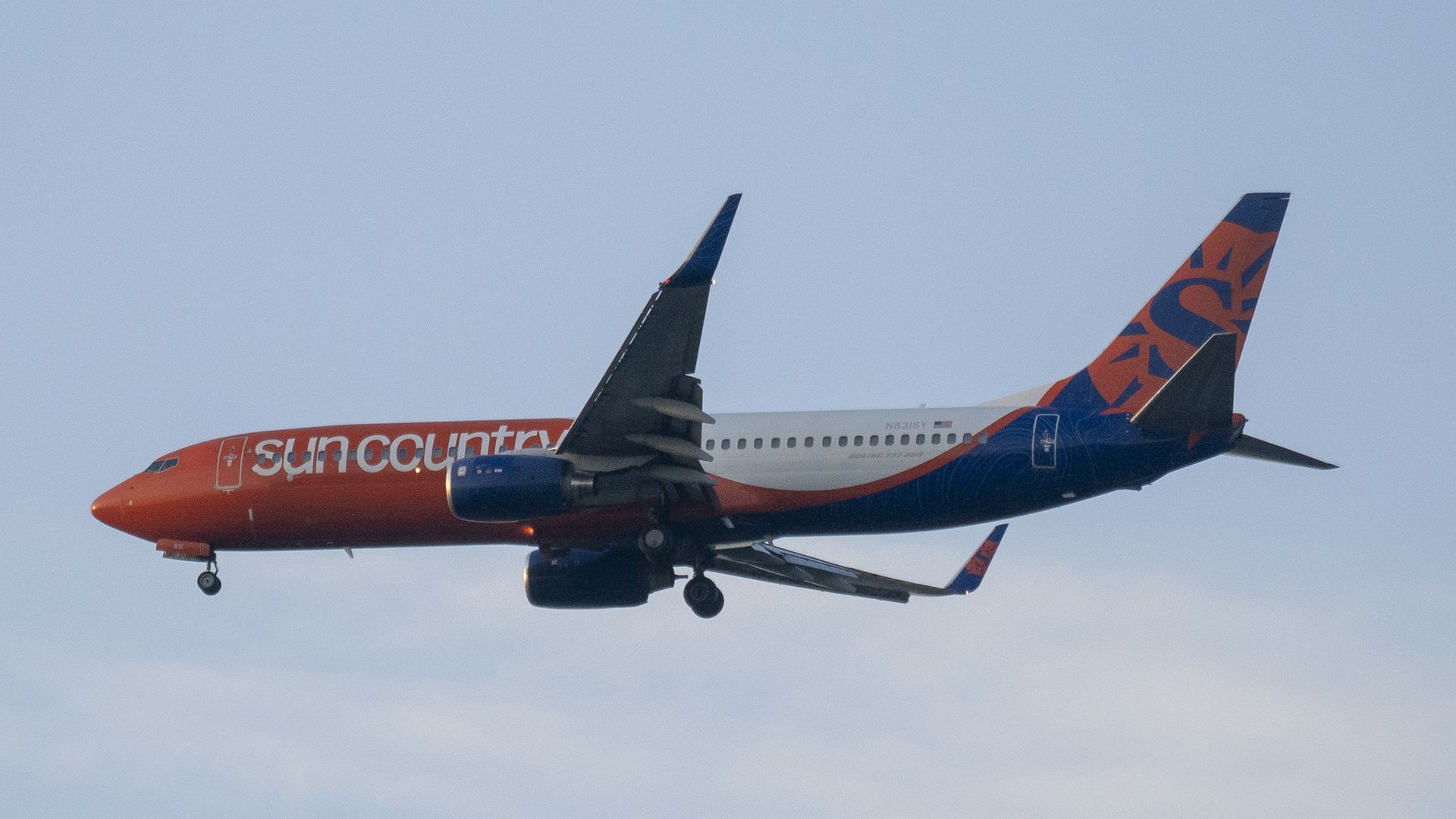 Sun Country Airlines airplane with orange front and blue tail featuring a sun logo, flying against a clear light blue sky.