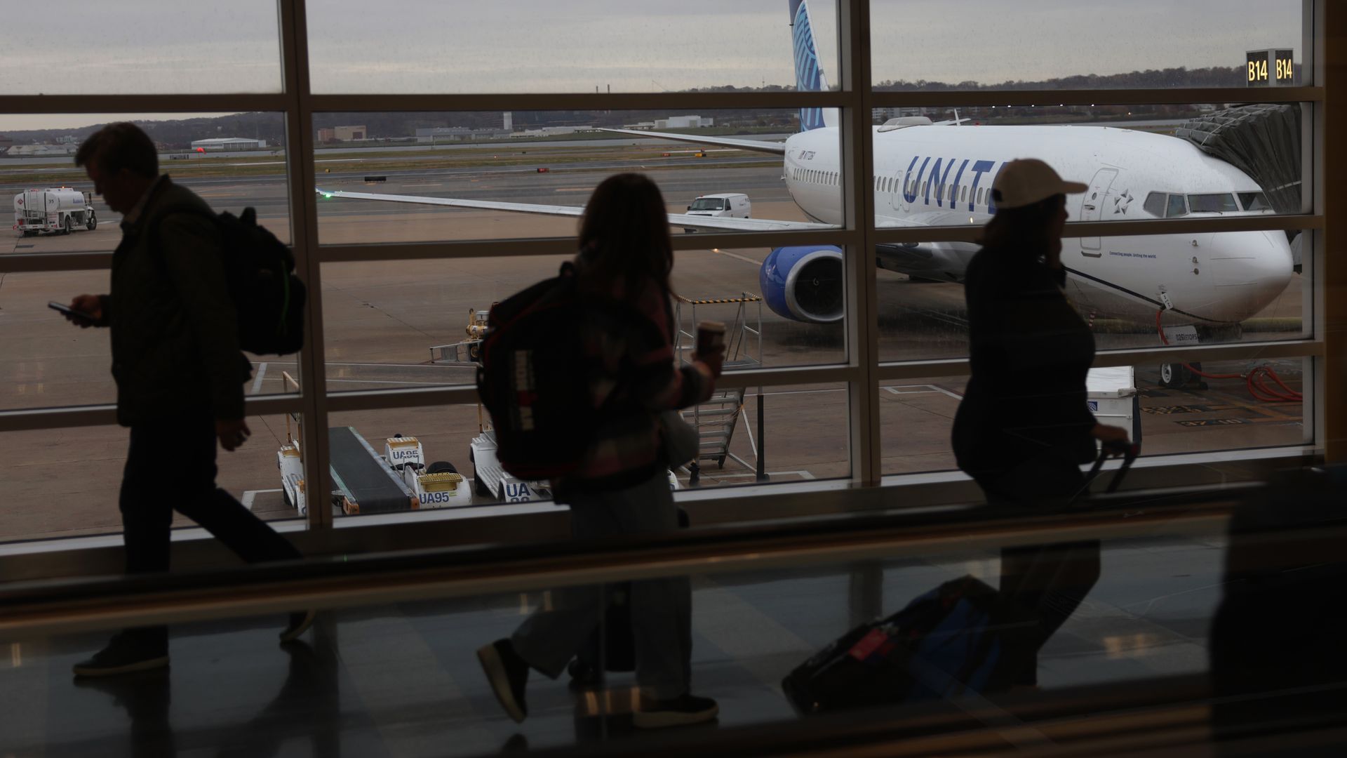 Passengers walk in front of a window in an airport, through which you can see a parked plane.