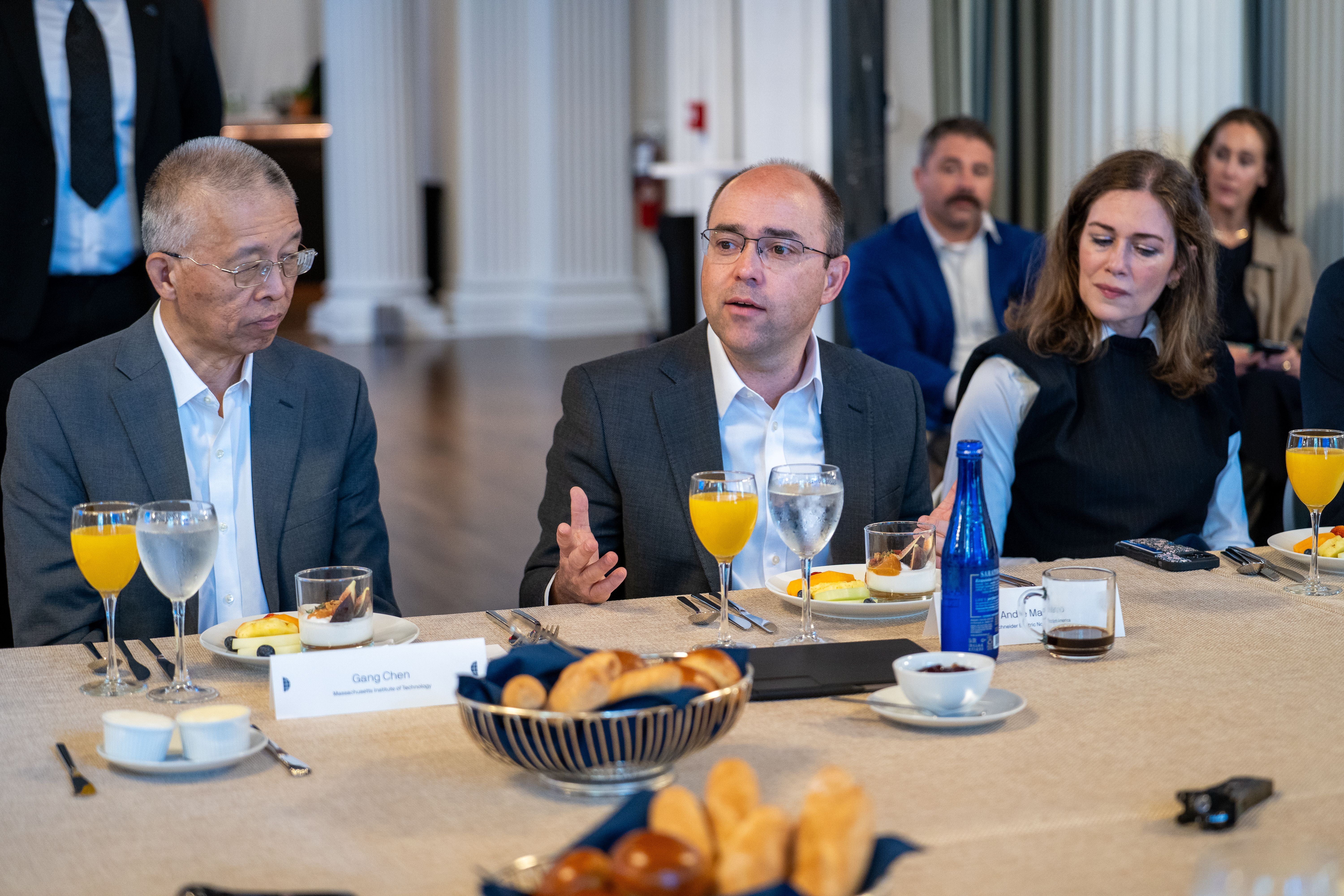 Business professionals at a formal dinner sit around a long table. The central man in a gray suit speaks, flanked by a glasses-wearing man and a woman with brown hair; table features drinks and bread.