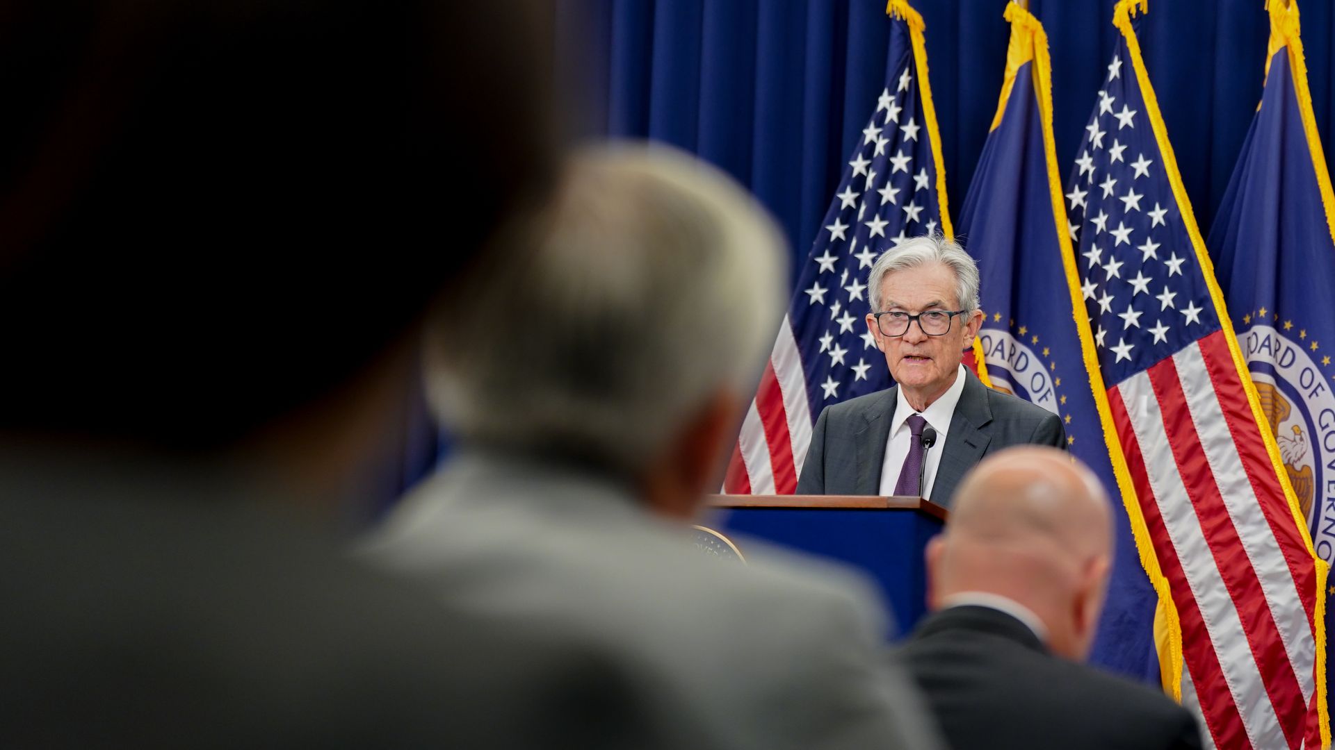 Fed chair Jerome Powell speaking at podium with microphone, American and official flags behind him, audience members blurred in foreground, blue curtain backdrop