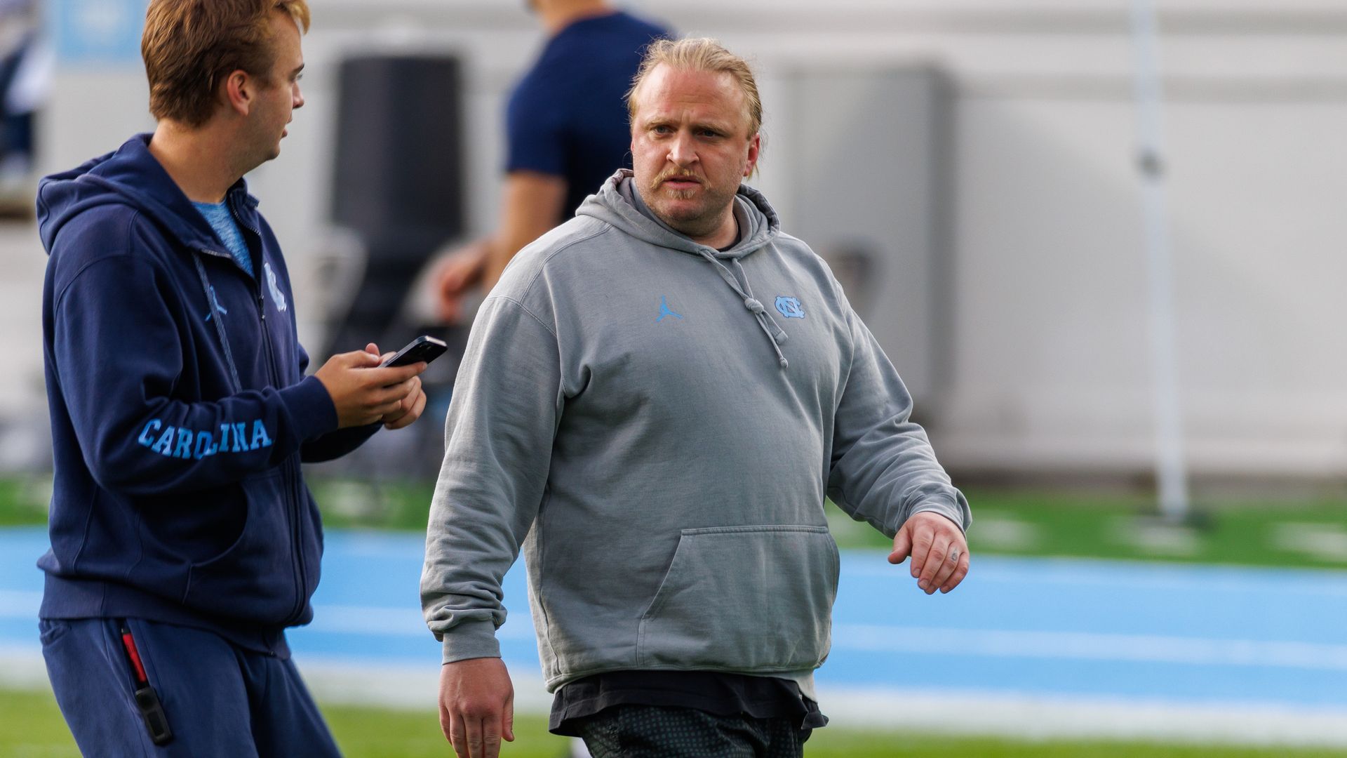 Two men on a sports field, one wearing a gray hoodie with a North Carolina logo and the other in a navy blue hoodie reading "Carolina," interacting with a phone in hand.