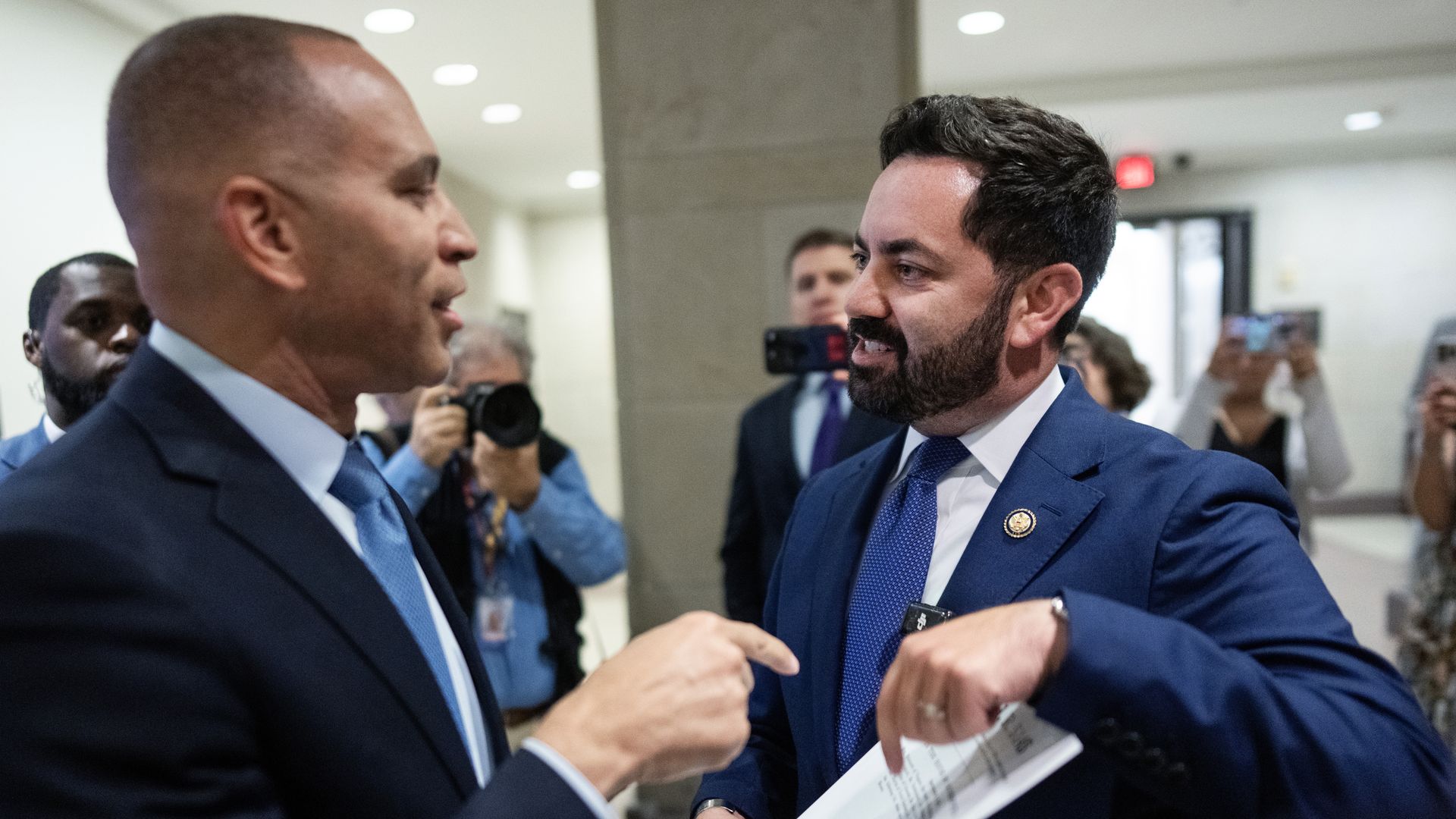 Hakeem Jeffries and Mike Lawler, both wearing blue suits, argue and point at each other while surrounded by press and photographers in a white and sandstone hallway.