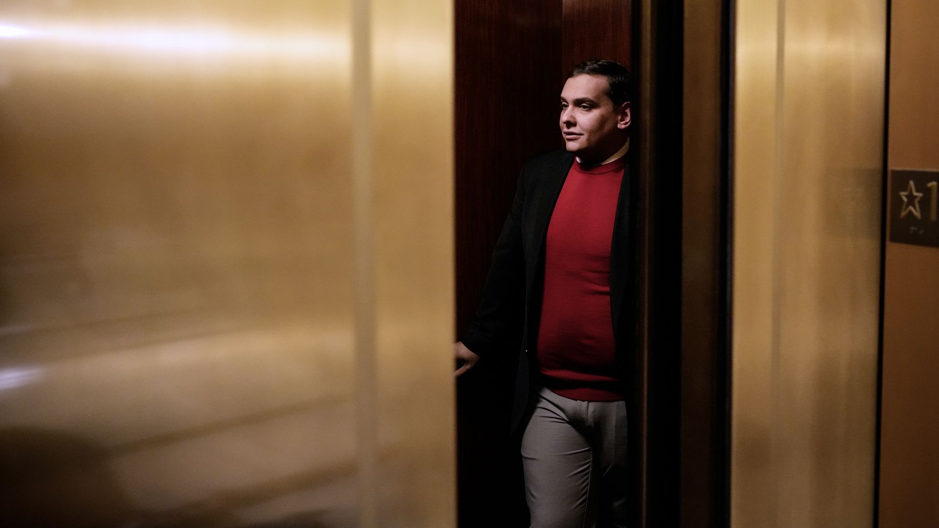 Santos, wearing a red sweater, behind closing elevator doors.