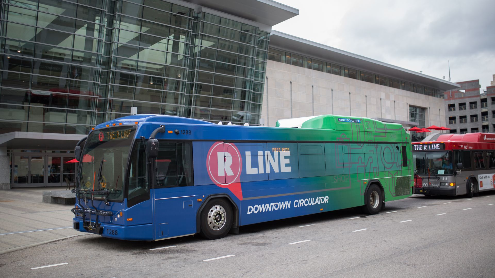 Blue and green R LINE bus labeled "Downtown Circulator" parked on a city street in front of a glass-fronted modern building; another red bus in background.