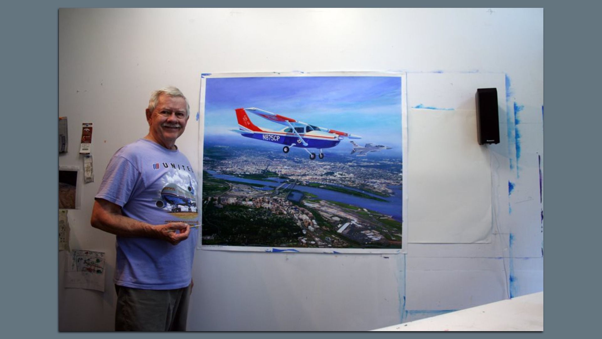 A man with silver hair and a purple shirt holds a paintbrush, standing to the left of a painting hanging on a white wall. The painting is of a prop plane, flying above a river landscape.