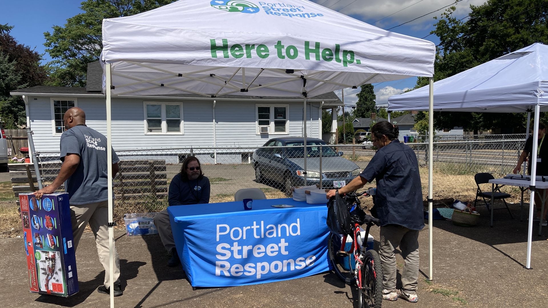 A man site at a table draped in a blue banner that reads Portland Street Response, under a white pop up tent that says Here to Help,  while a man with a bike reaches into. a cooler full of water bottles.