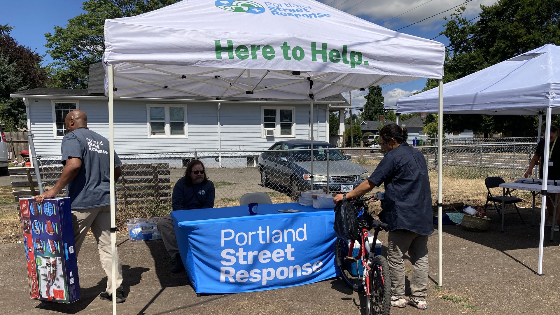 A man site at a table draped in a blue banner that reads Portland Street Response, under a white pop up tent that says Here to Help, while a man with a bike reaches into. a cooler full of water bottles.