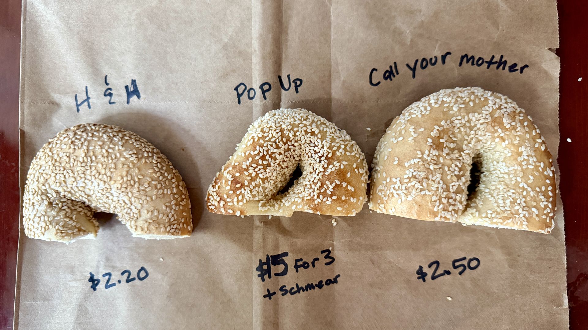 Three sesame-seed bagels on brown parchment with handwritten notes in black ink, including "POP UP" and "Call your mother," with faint price scribbles nearby.