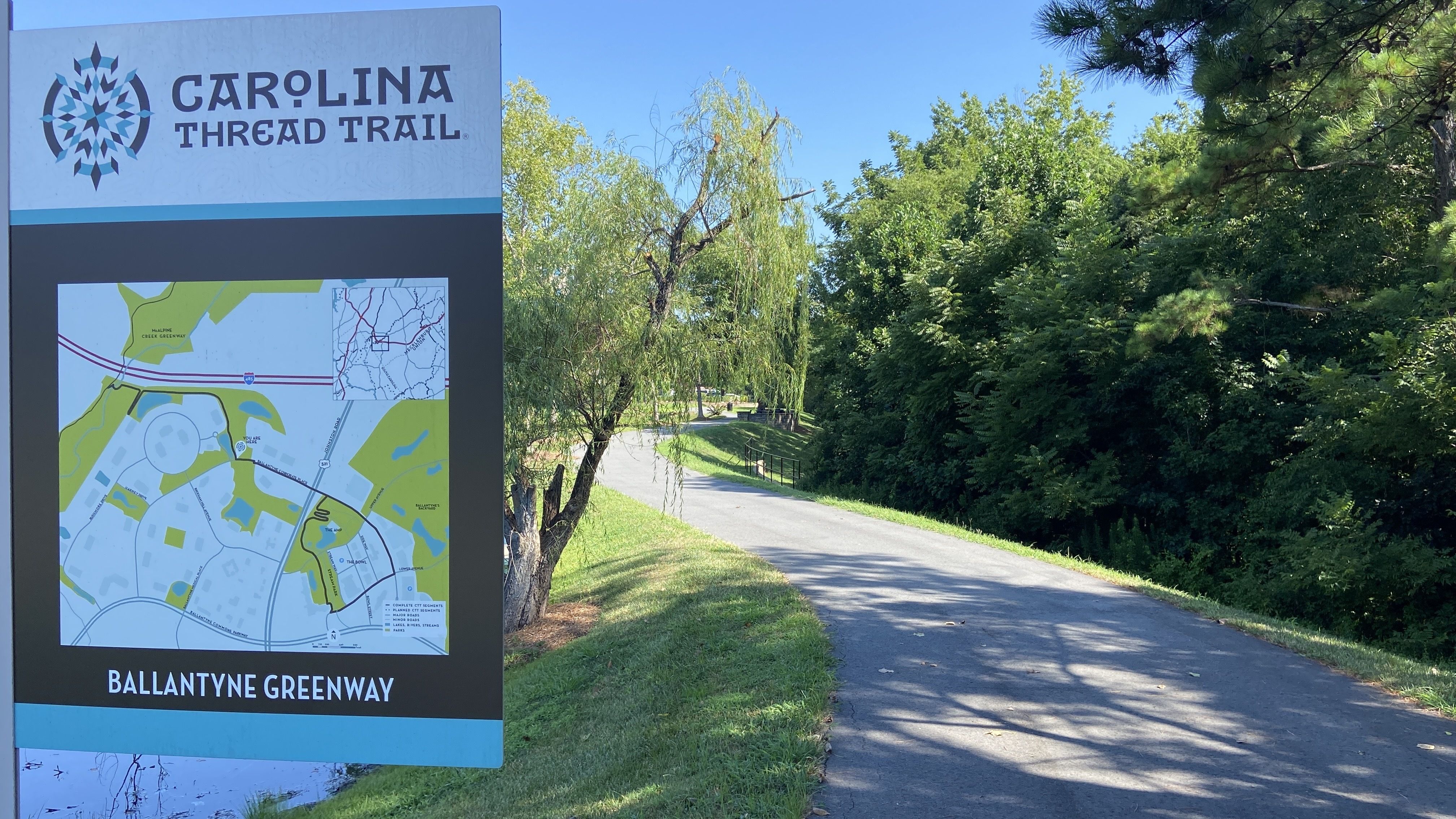 Sign for Carolina Thread Trail at Ballantyne Greenway beside a paved path with green grass, trees, and clear blue sky on a sunny day.