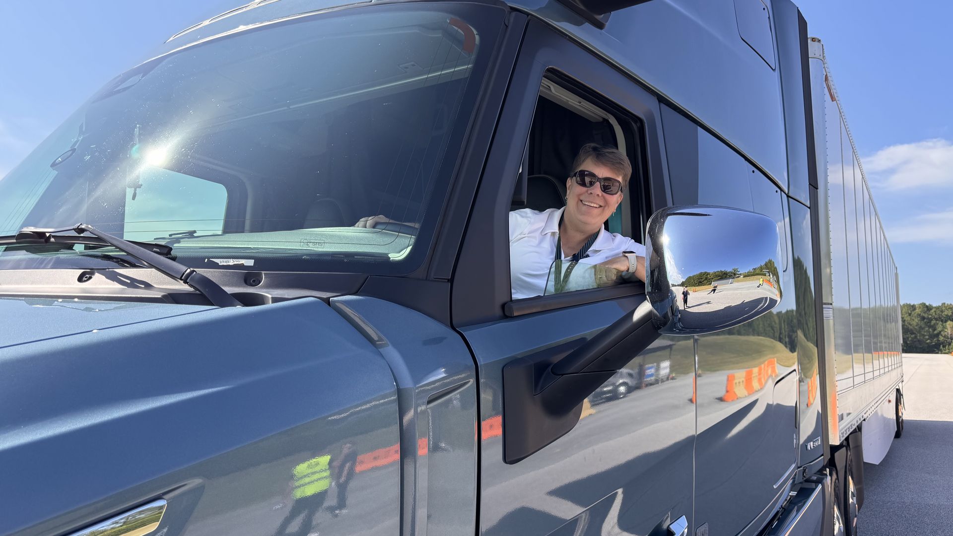 Author Joann Muller wearing sunglasses and white shirt smiling while leaning out window of a dark blue Volvo semi truck on a sunny day with clear blue sky.