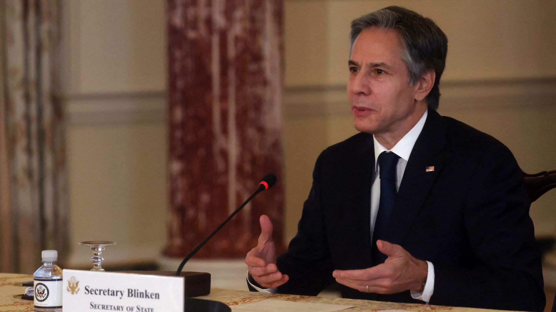 Blinken wears a suit and tie while sitting in front of a microphone and a placard with his name on it