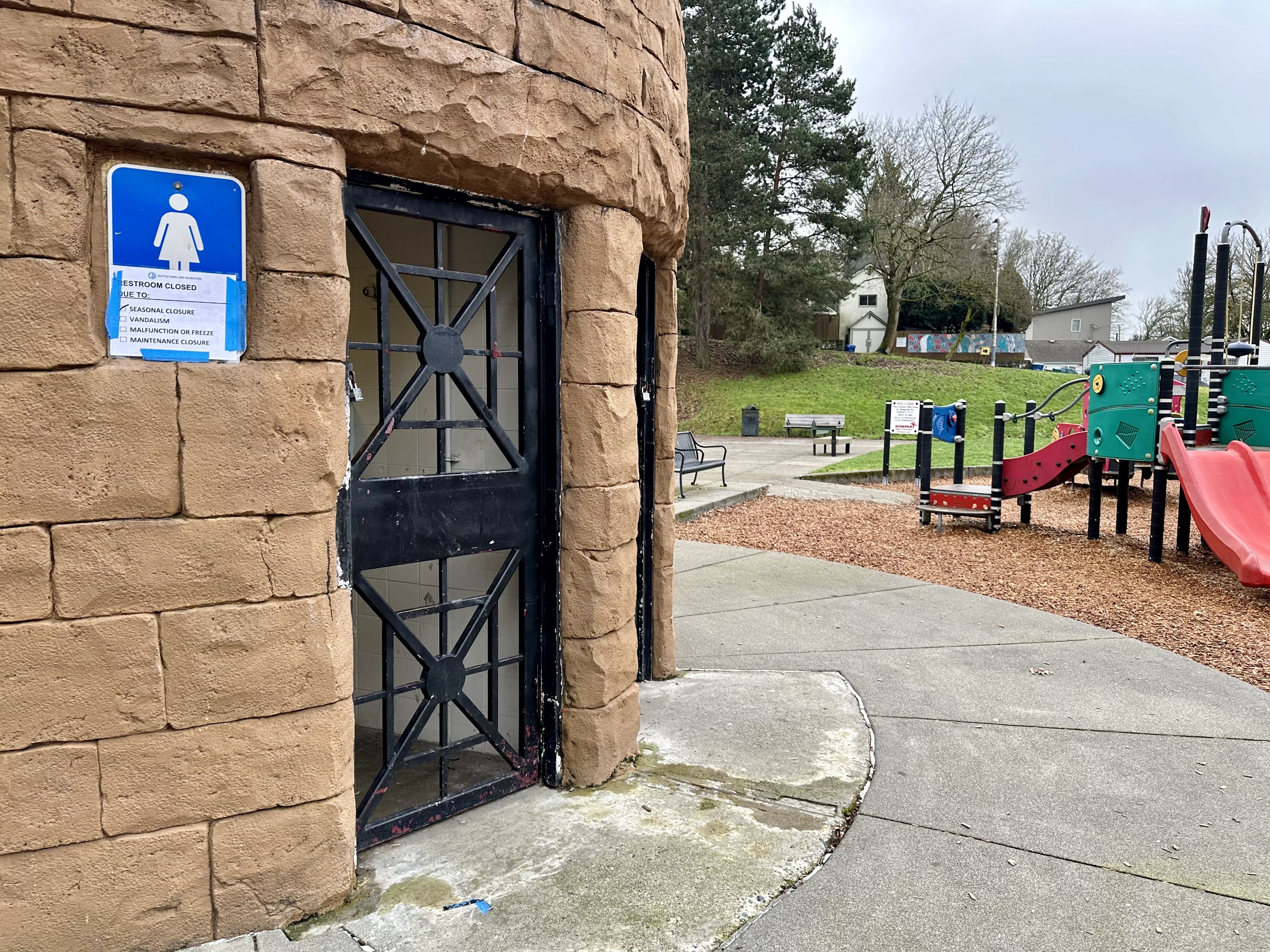 A standalone bathroom building locked up with a sign over the women's sign that says closed for the season, next to a children's play area.
