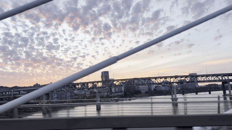 Animate gif shows the view from a moving train going over a bridge, with high clouds illuminated at sunset over the downtown skyline of Portland.