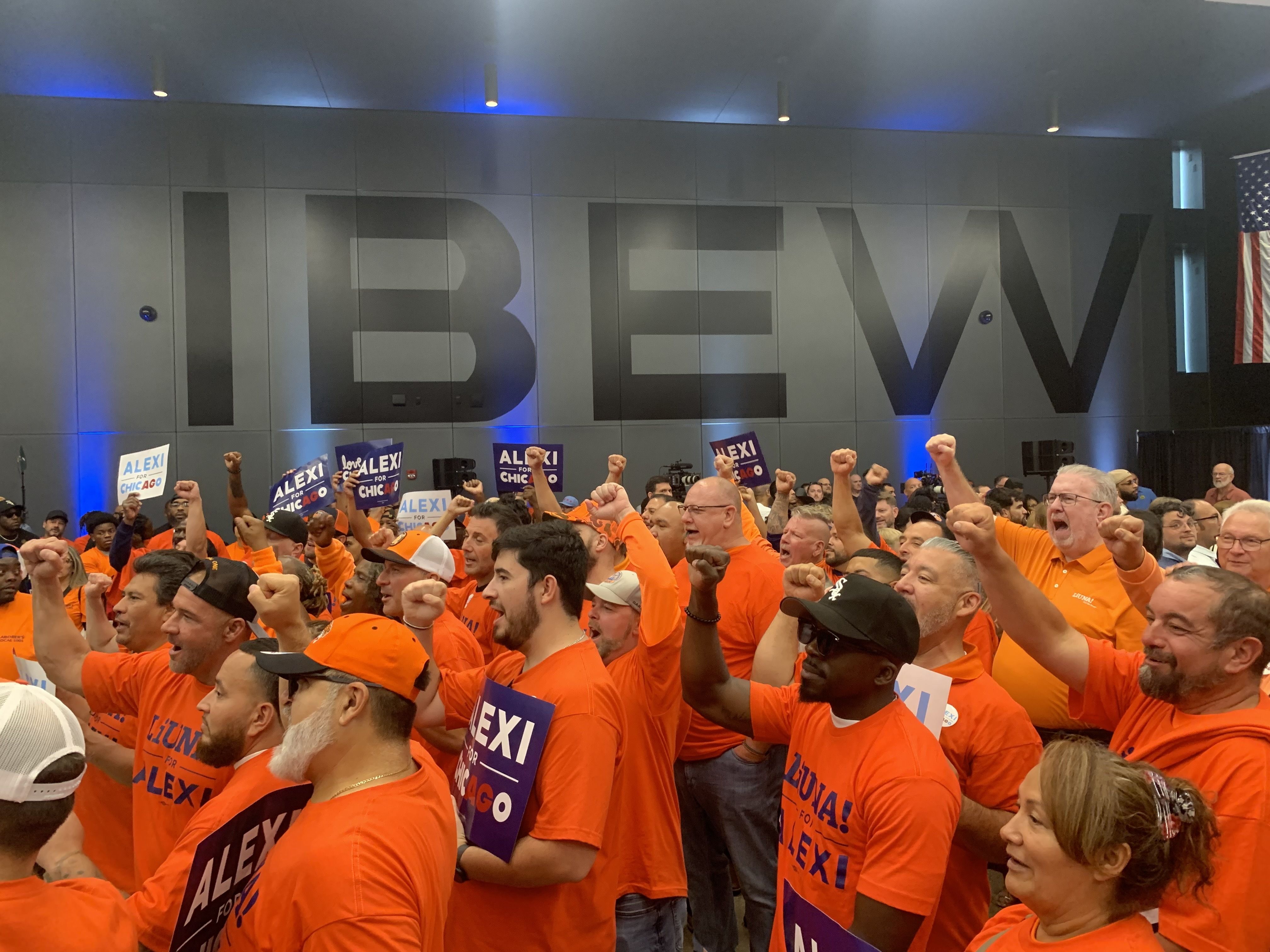 Indoor political rally with a sea of people in bright orange shirts, many holding blue signs reading "ALEXI FOR CHICAGO". Blue lights along a gray wall and an American flag on the right.