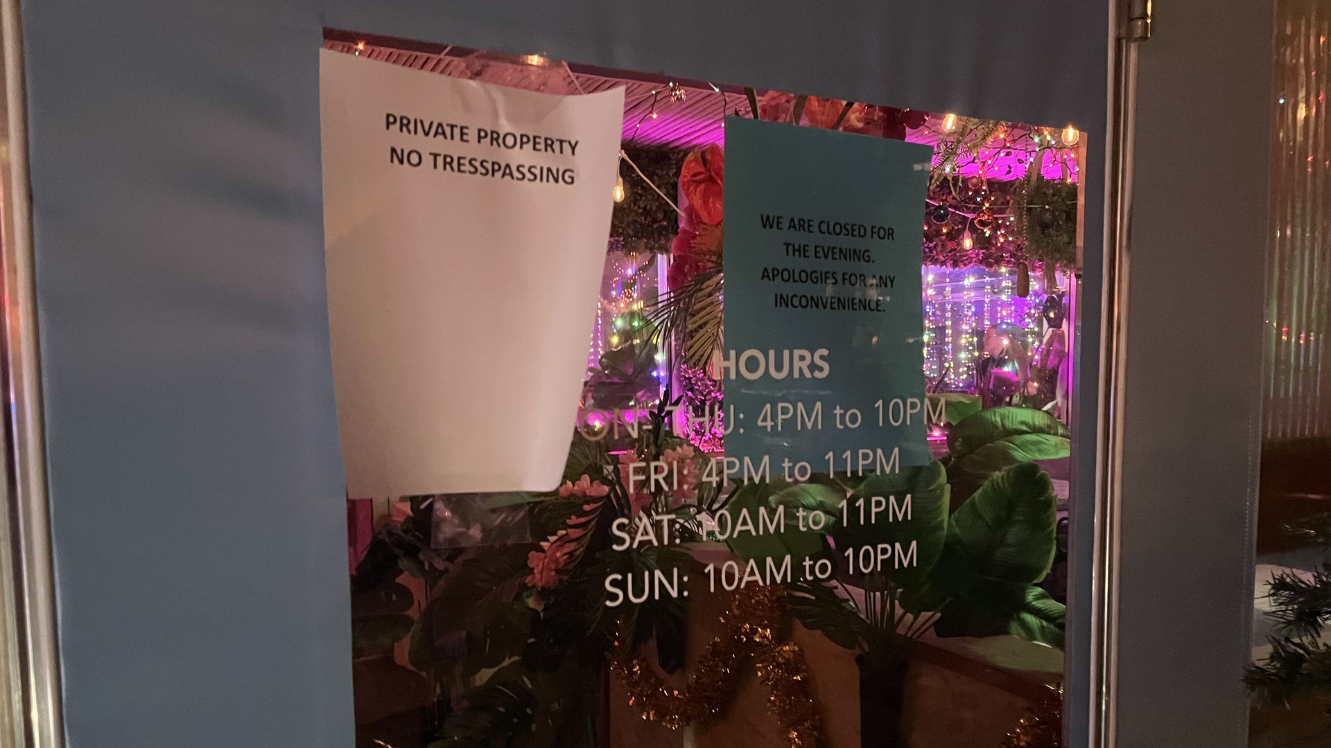 Blue door with reflective window showing colorful lights inside. Signs say "Private Property No Trespassing" and "We are closed for the evening. Apologies for any inconvenience." Hours listed.
