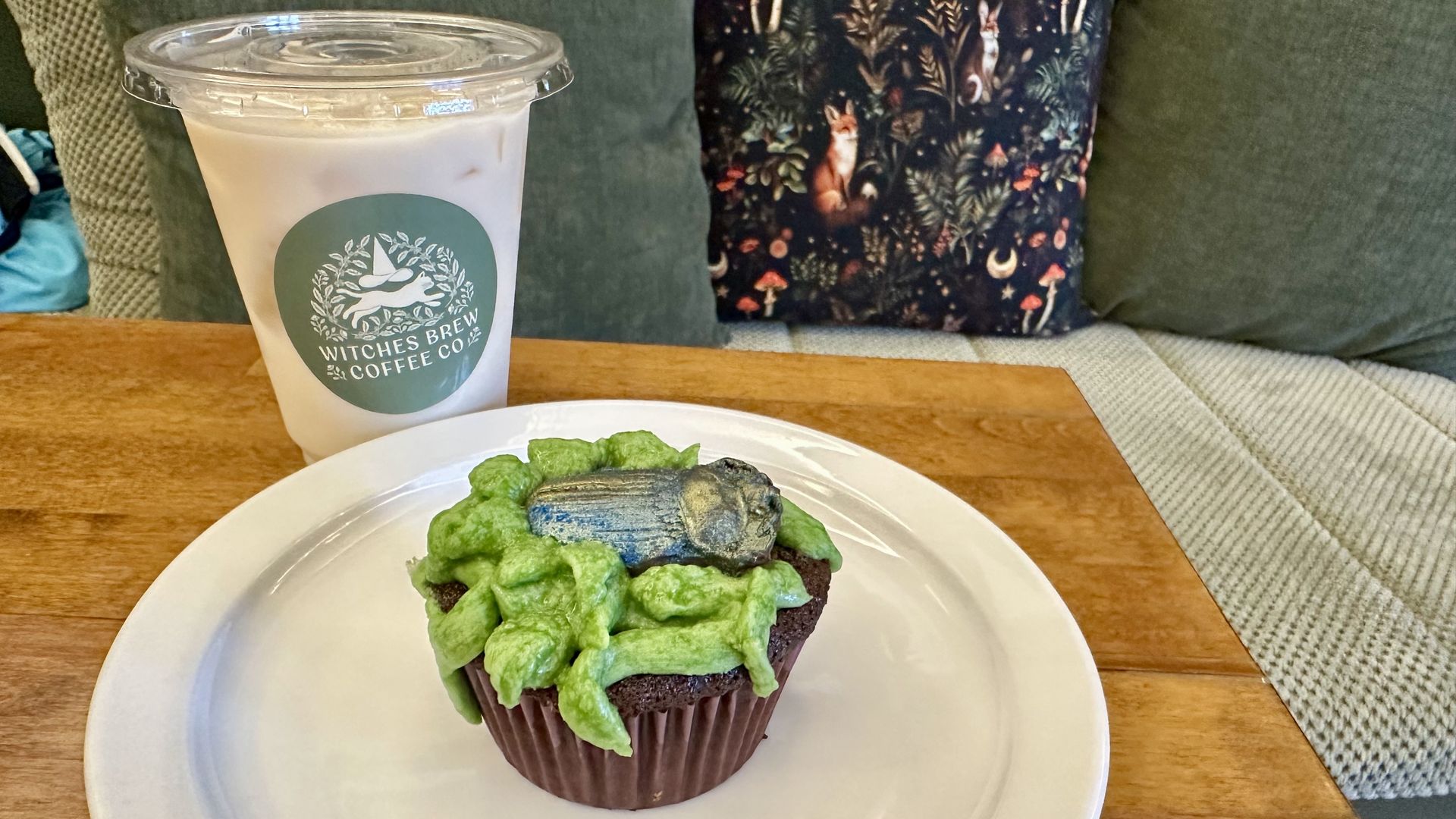 To-go iced coffee in a clear cup with a green Witches Brew Coffee Co. logo sits on a wooden table beside a plate with a green-frosted cupcake.
