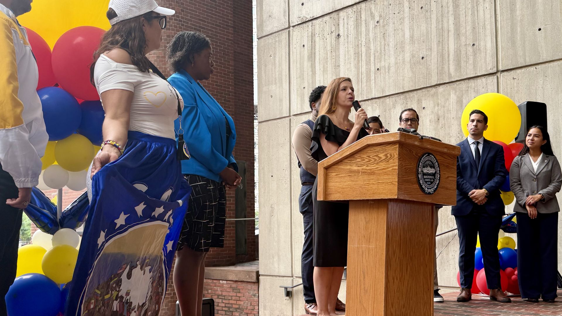 Carlina Velásquez (left), president of the Venezuelan Association of Massachusetts, holds her full-length blue skirt to show a mural of Venezuelan as Cristina Aguilara Sandoval (right) speaks at a lectern at Boston City Hall. Sandoval leads the Mass. Office for Refugees and Immigrants.