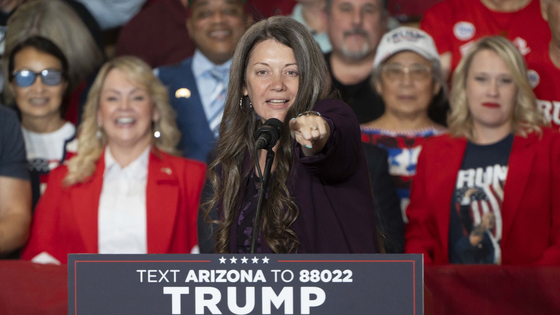 A woman with long brown hair points forward at the camera while standing at a lectern and a microphone with a sign in front of it that reads "Trump Vance." 