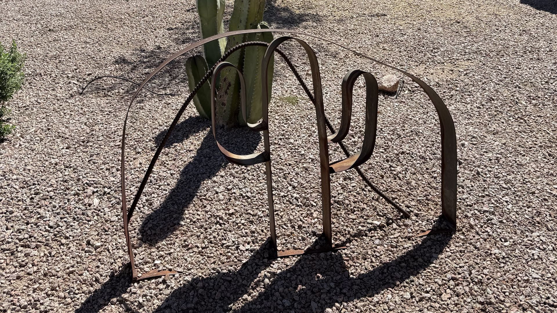 Broad, rust-colored metal strips shaped into a two-armed saguaro cactus, with another strip forming an outline around it; the sculpture is on a gravel field with cactus behind it.