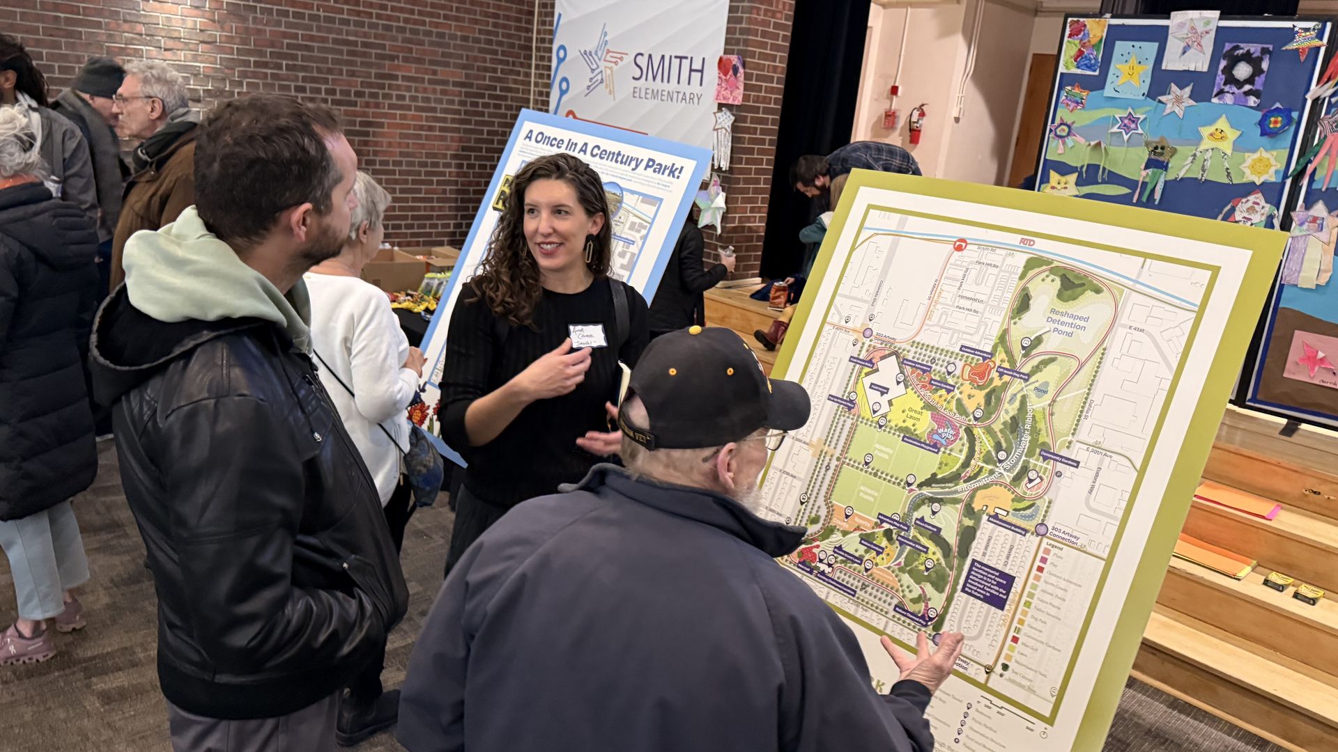 People gathered indoors at Smith Elementary, discussing a large park plan display and a poster titled "A Once In A Century Park!" with a brick wall and colorful children's artwork in background.