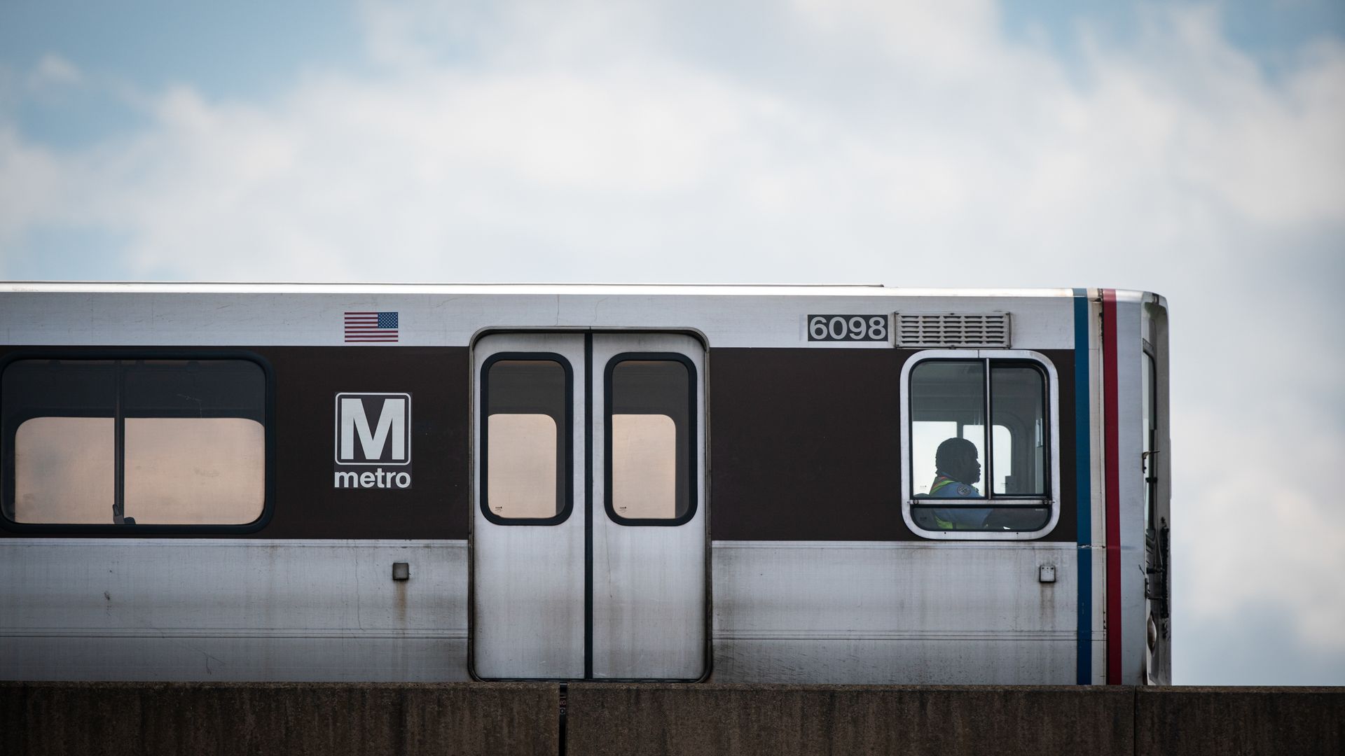 Side view of a silver and brown Metro train car number 6098 with an American flag decal, a closed door, and a silhouette of a person inside against a blue sky with clouds.