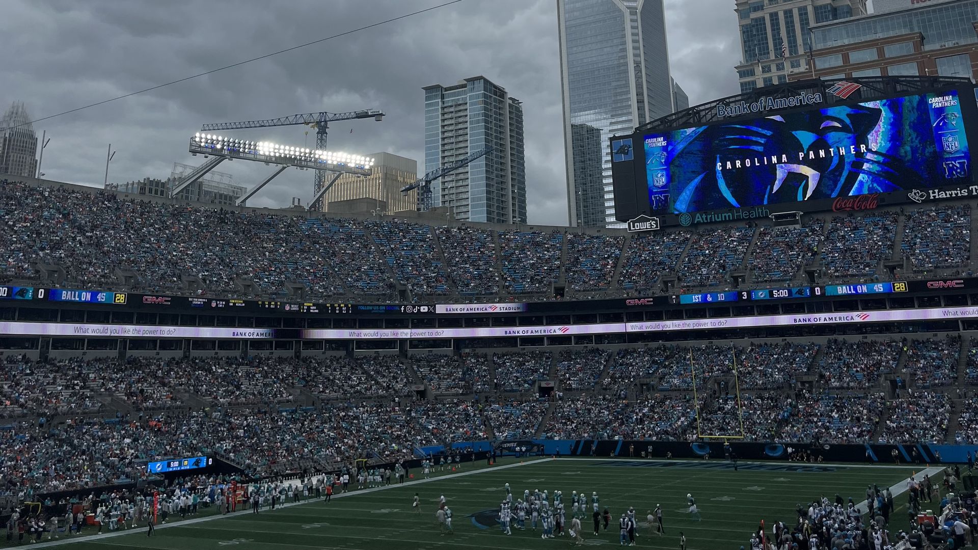 View of a crowded Carolina Panthers football stadium under gray clouds with players on the field and a large scoreboard showing the Panthers logo and game info.