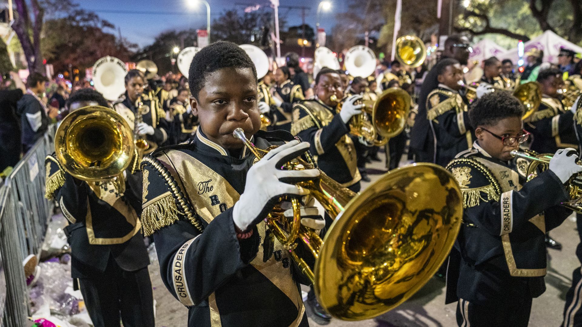 A marching band performs music as they march along a parade route. The camera is focused on a young trombone player.