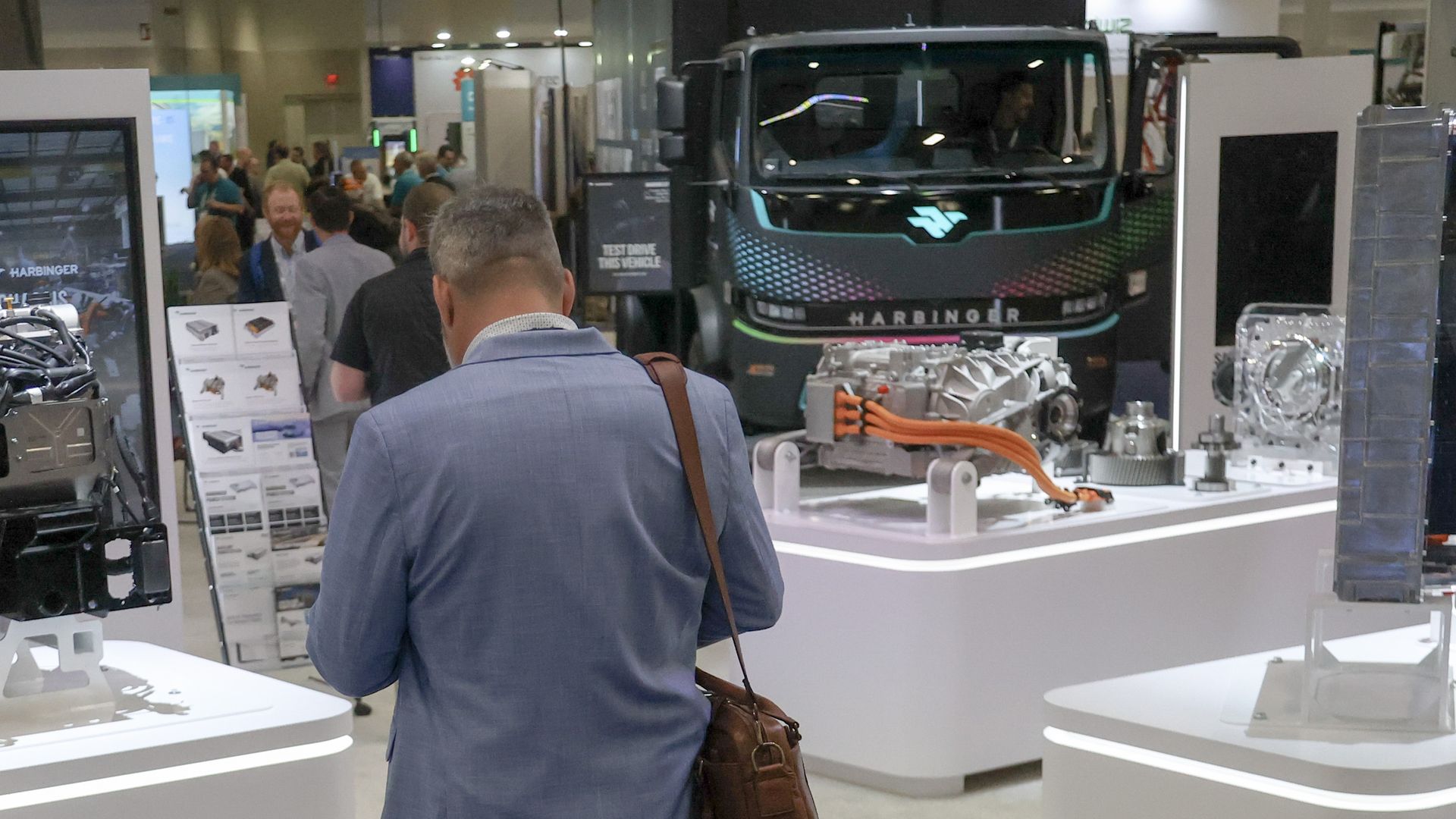 A photo of a conference attendee in a light-blue sportcoat, apparently looking at his phone while facing away from the camera, with an electric Harbinger Motors panel truck parked in the background of the conference floor.