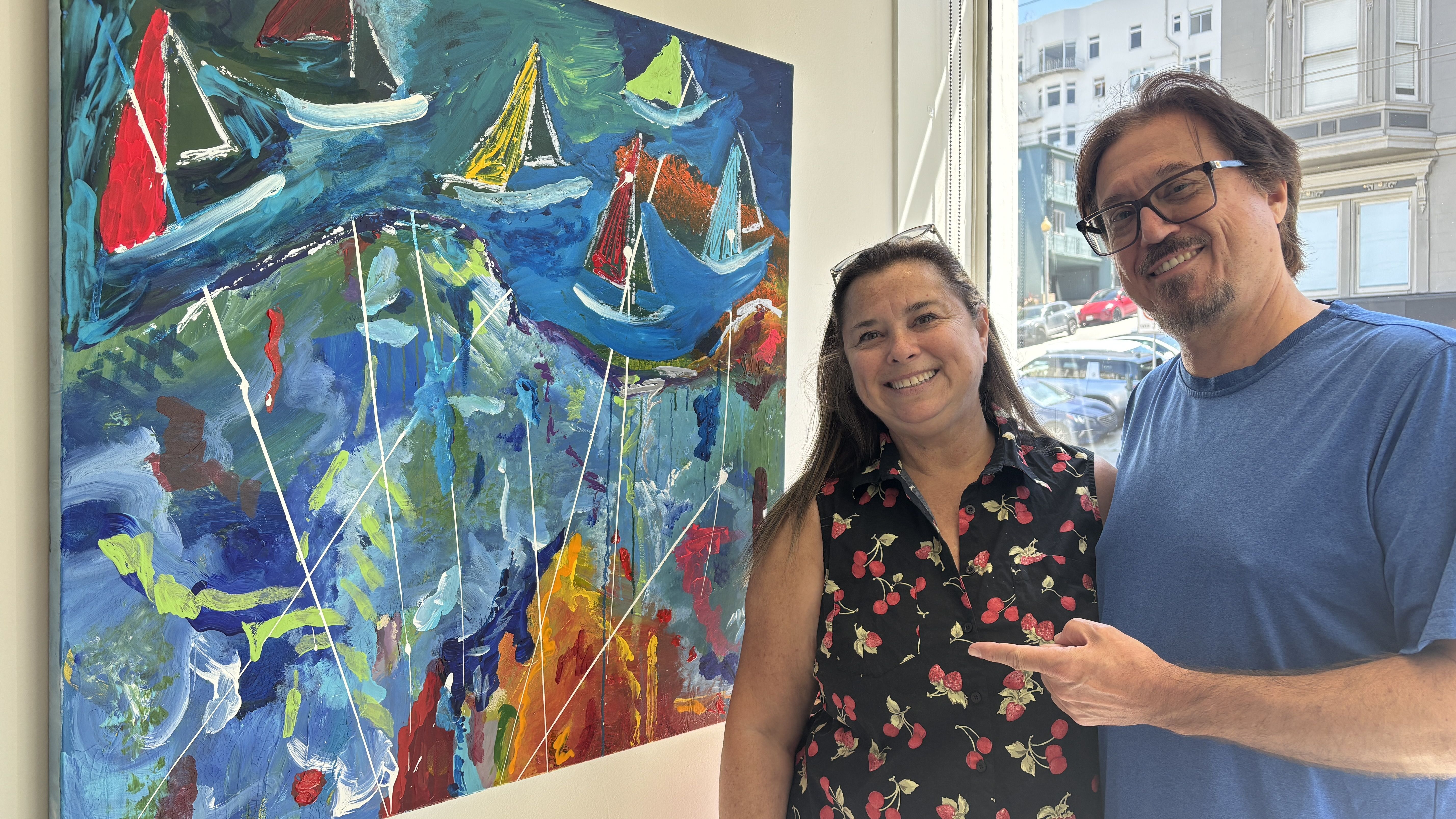 Smiling woman in a floral blouse and a man in a blue T-shirt stand beside a large, colorful abstract painting of sailboats with white mast lines; city street visible through the window.