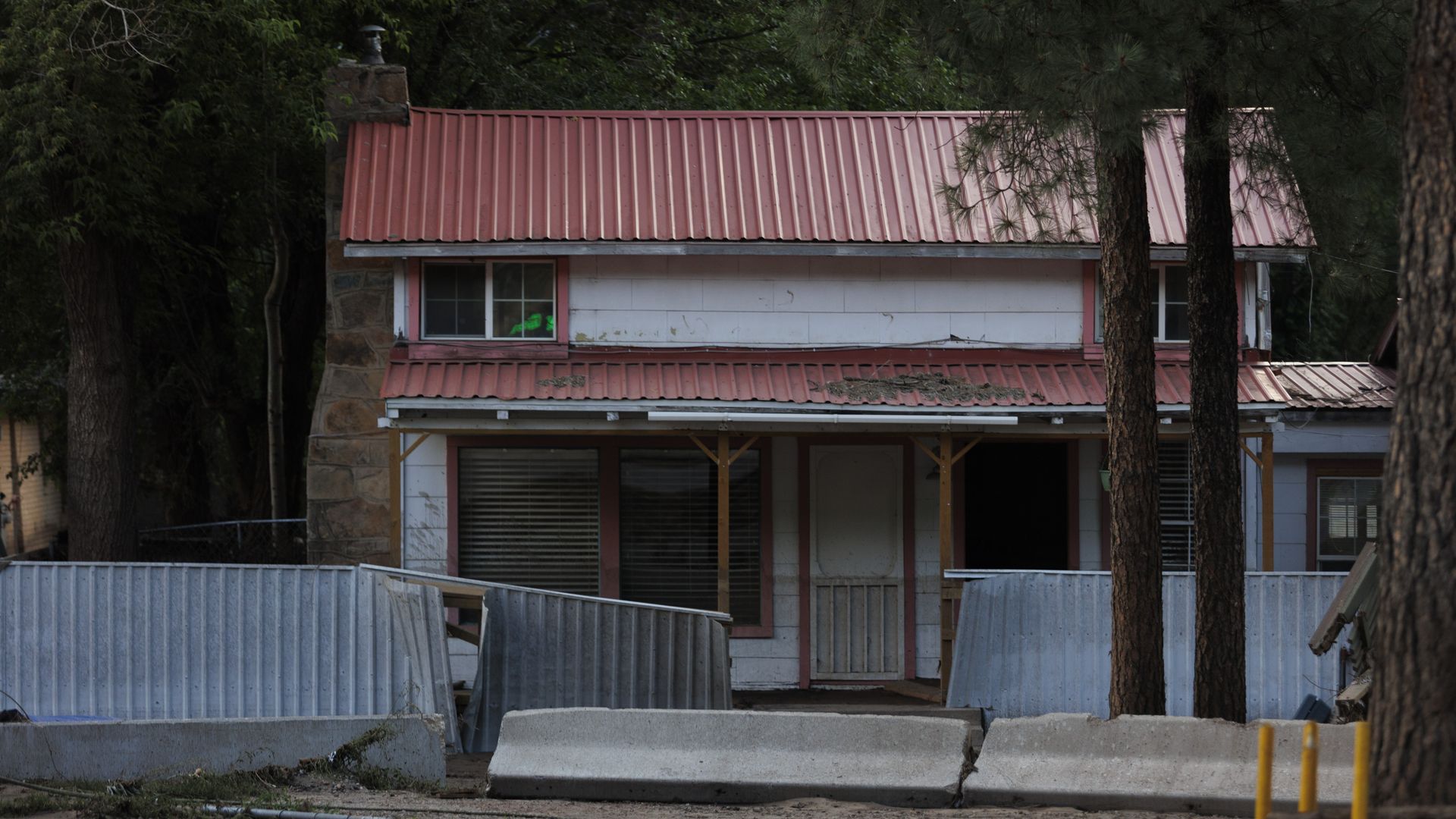 Old two-story house with a red metal roof, white walls, and boarded windows behind fallen metal fences and concrete barriers, surrounded by tall trees in a forested area.