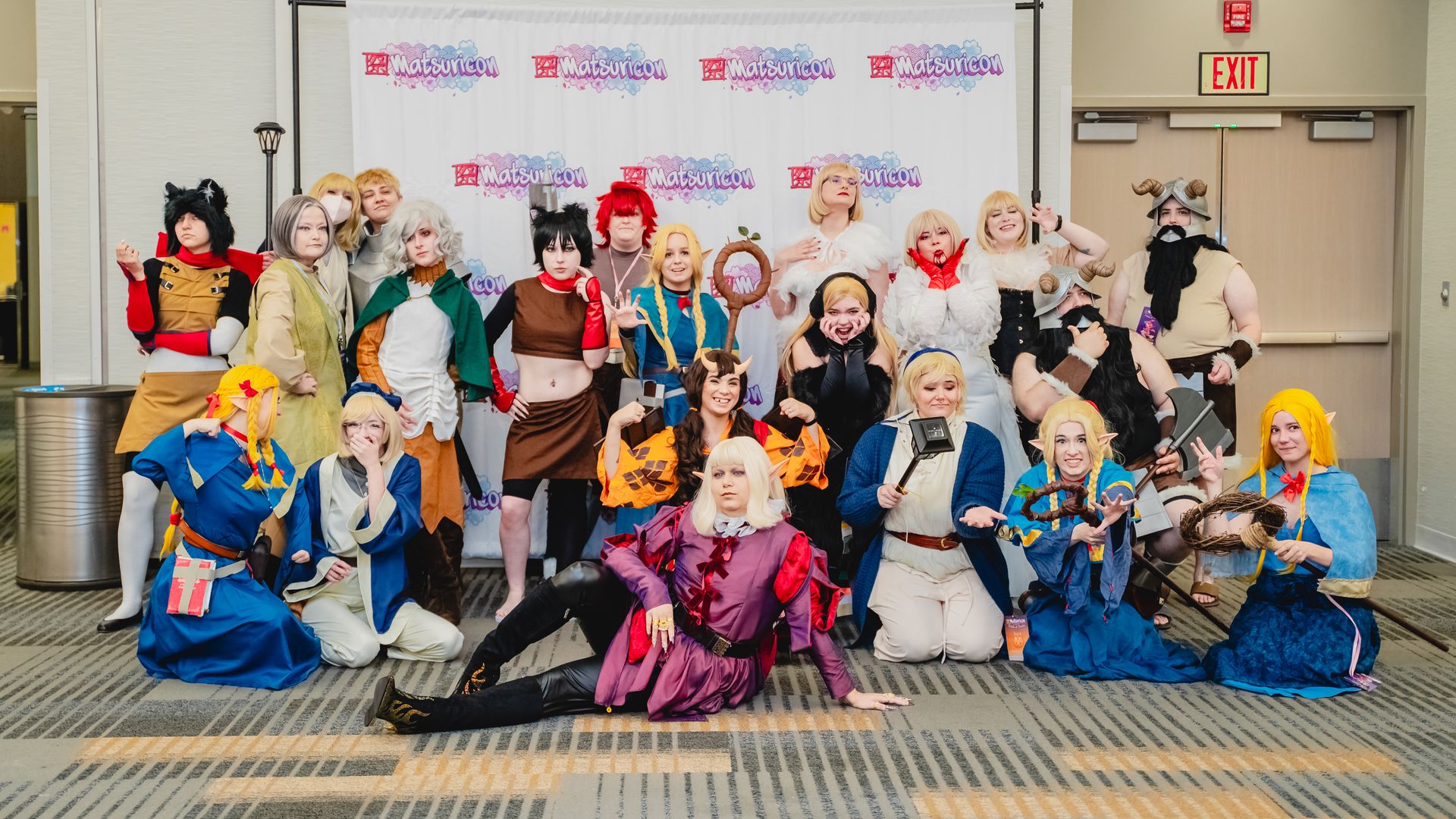 Group of 20 cosplayers in diverse colorful costumes posing inside a convention center, standing or crouching in front of a white "Matsuricon" backdrop banner.