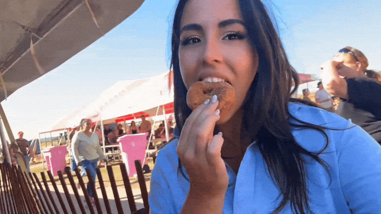 A woman with dark hair and a blue shirt eats a powdered donut at an outdoor event with tents, fencing, and people in the background under a clear blue sky.