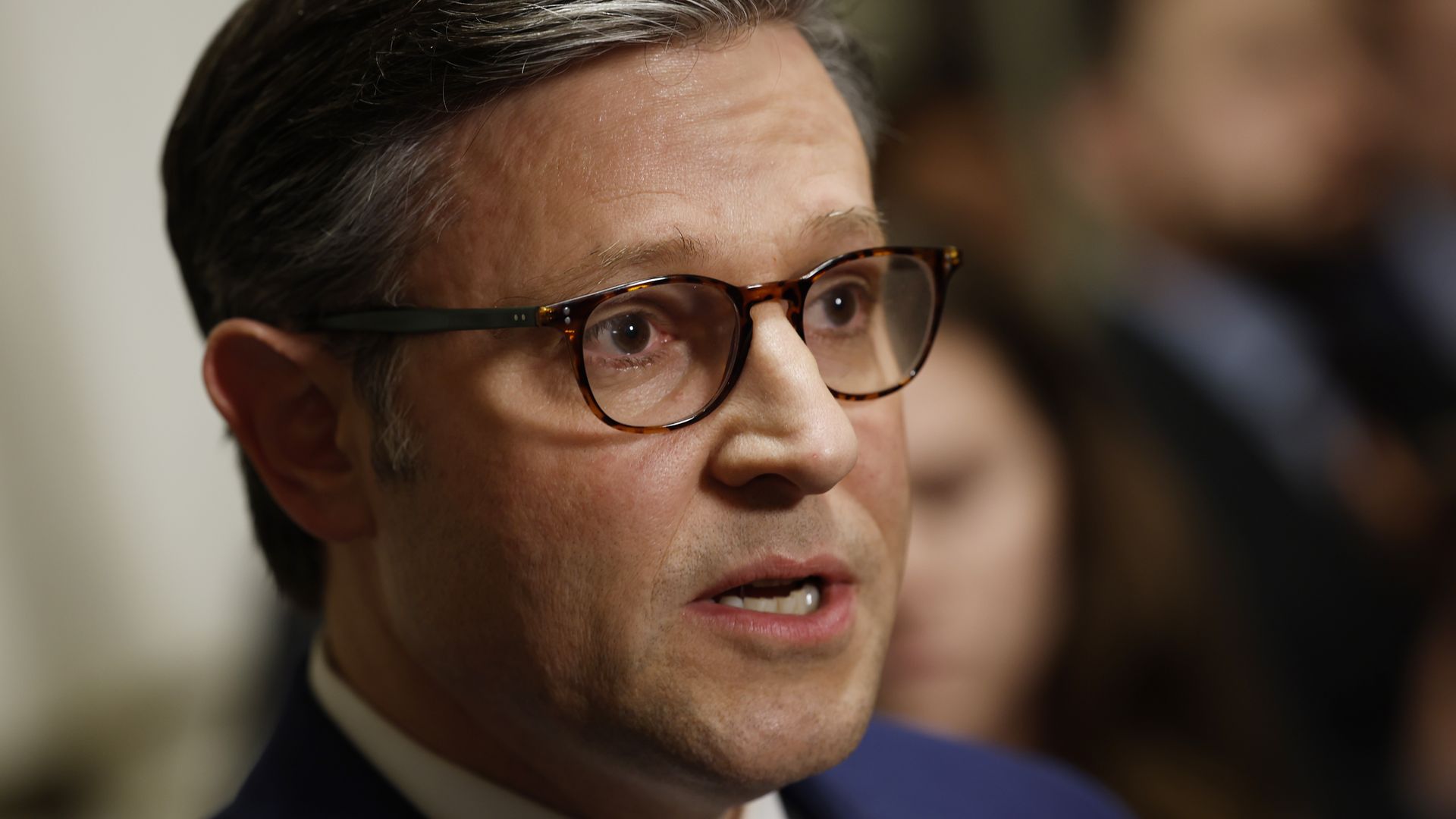 House Speaker Mike Johnson, wearing a blue suit and glasses and surrounded by reporters.