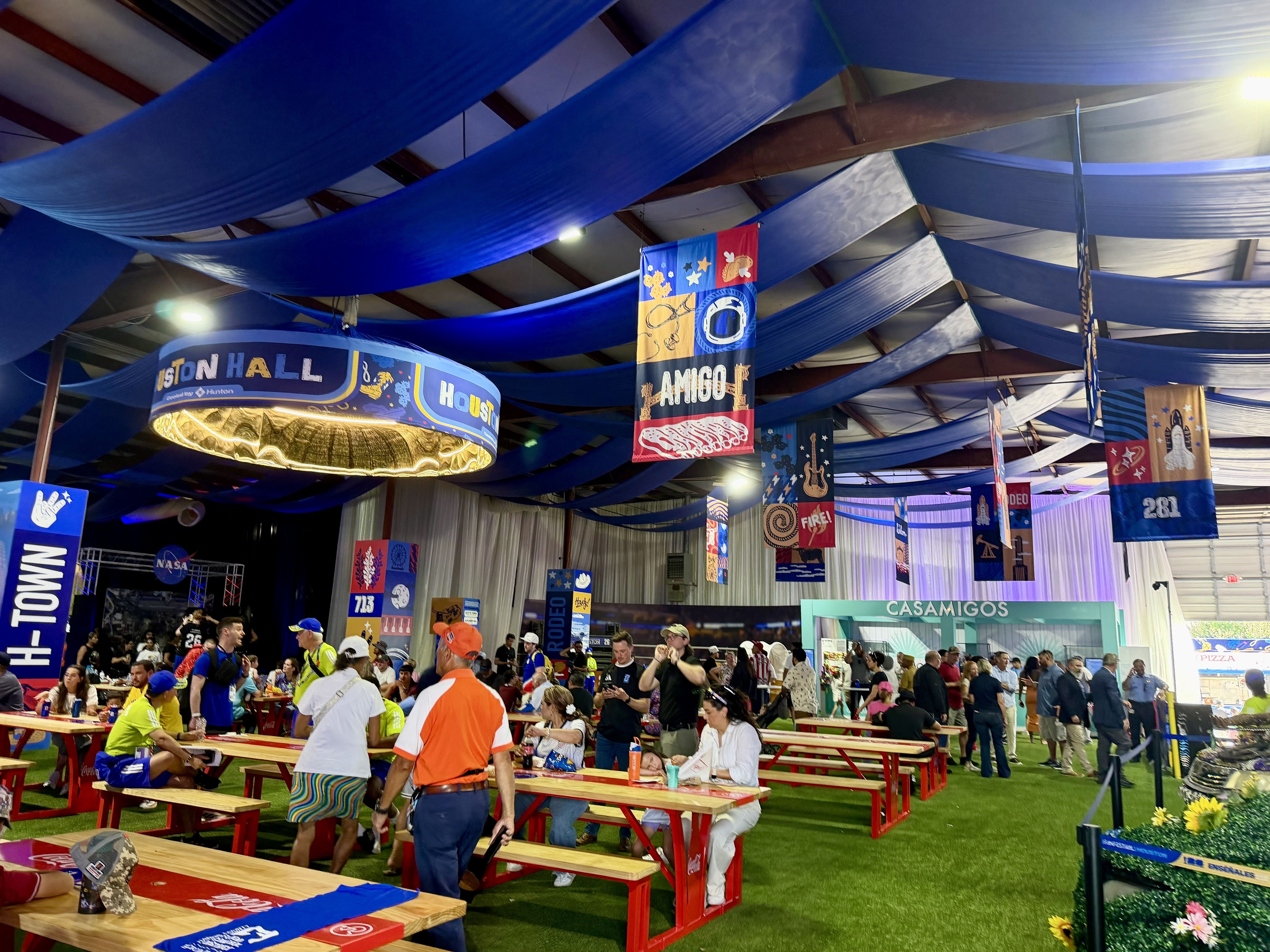 A large indoor event hall with blue fabric draped across the ceiling, colorful banners hanging down, and crowds seated at red picnic tables eating; green carpeted floor and bright lights.