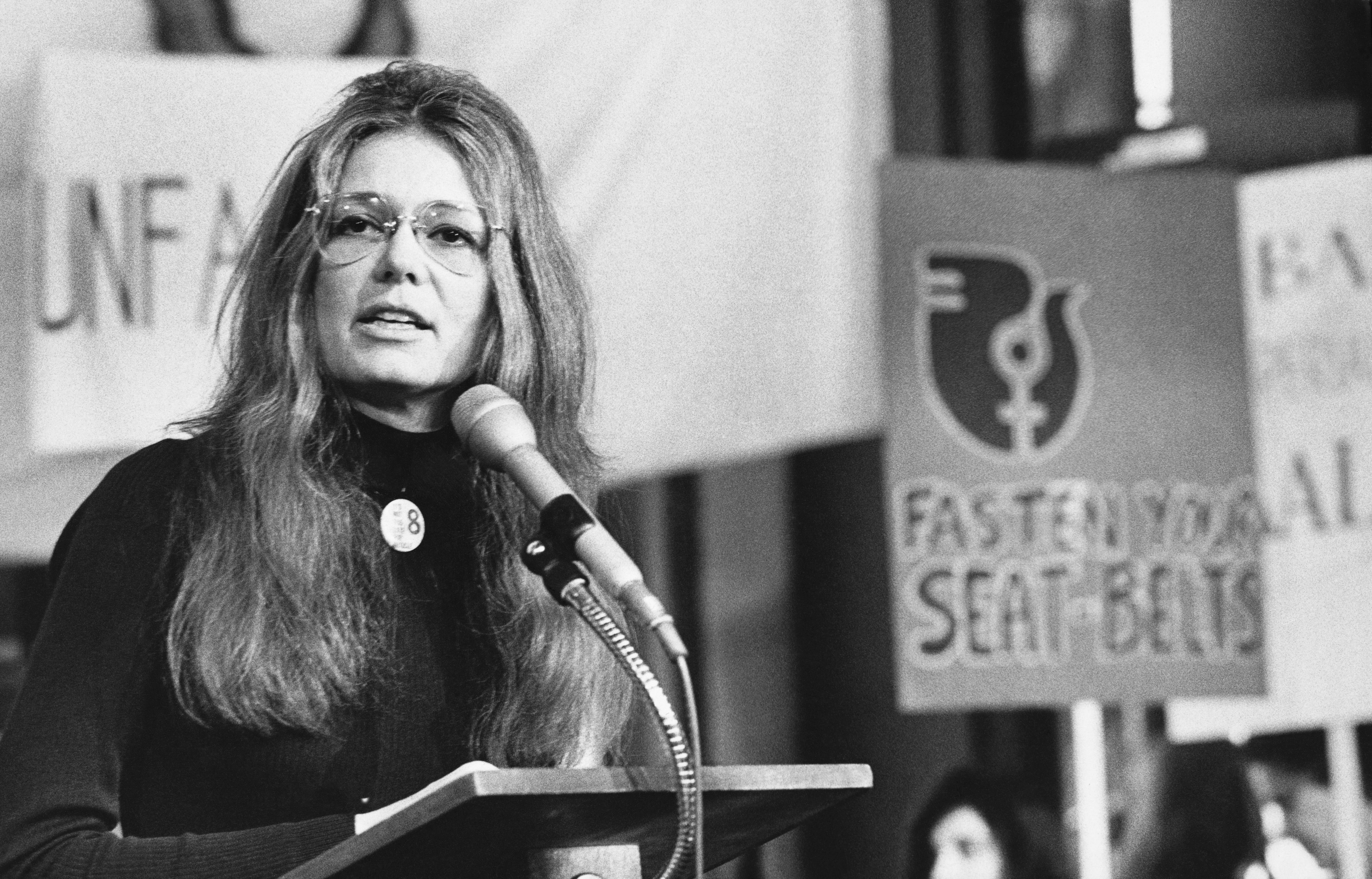 Gloria Steinem speaks into a microphone attached from behind a podium at a rally while wearing a turtleneck and glasses.