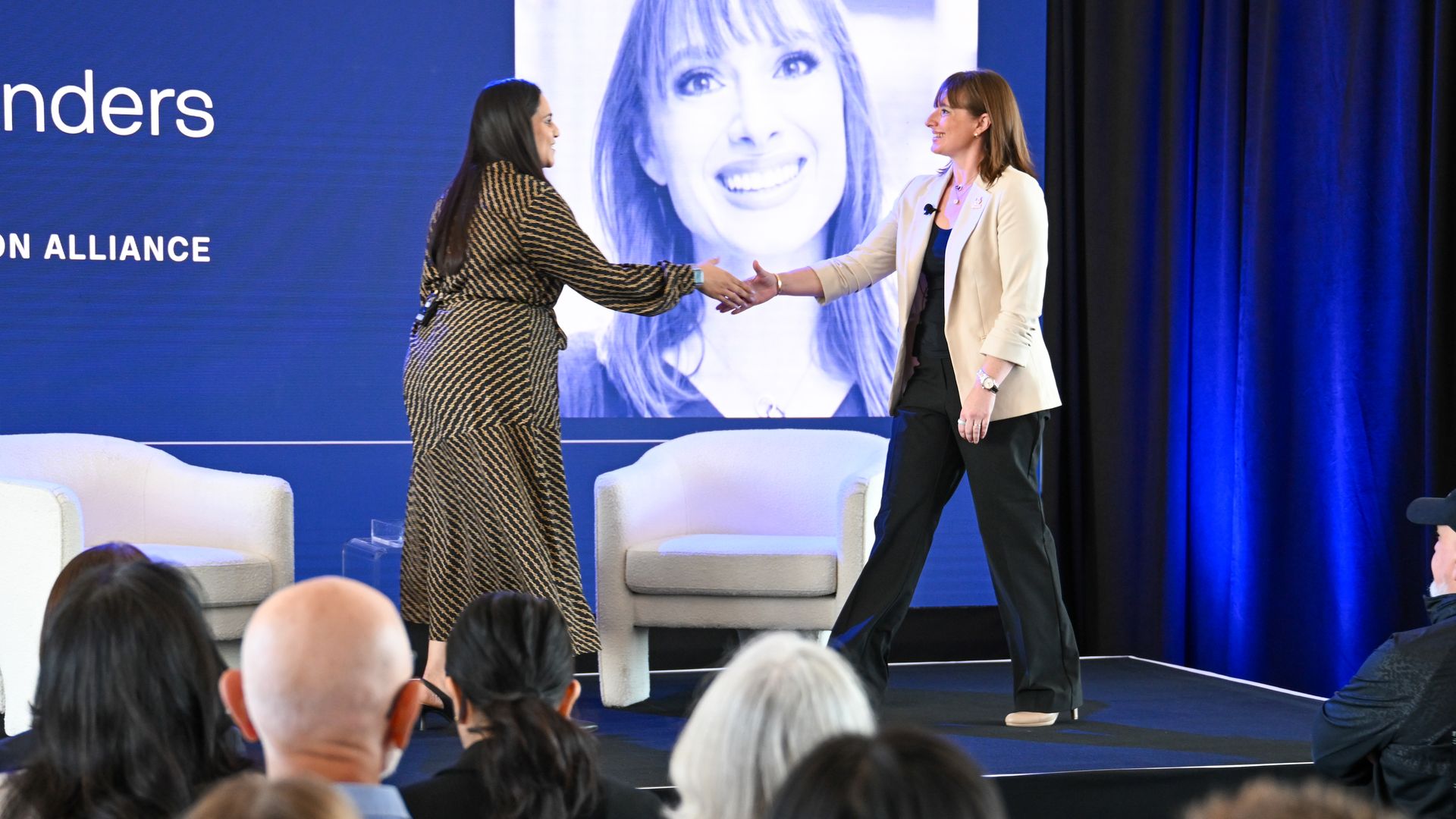 Two women shake hands on a conference stage. Left wears a patterned brown dress; right a beige blazer with black pants. A blue backdrop shows a large smiling portrait and the audience.