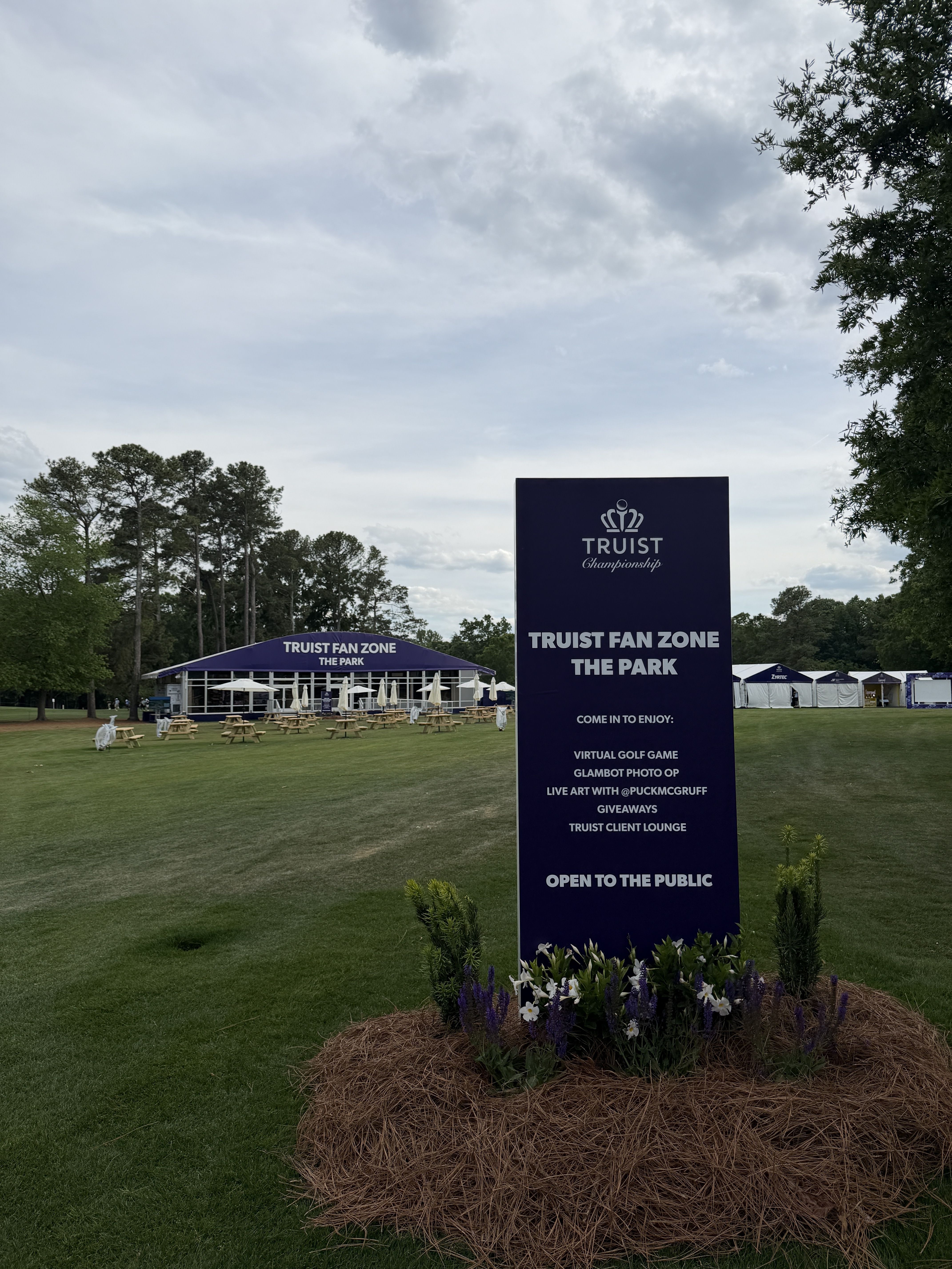 Outdoor park scene with a tall purple sign reading TRUIST FAN ZONE THE PARK in the foreground; blue tents and picnic tables with white umbrellas on a green lawn under a cloudy sky.