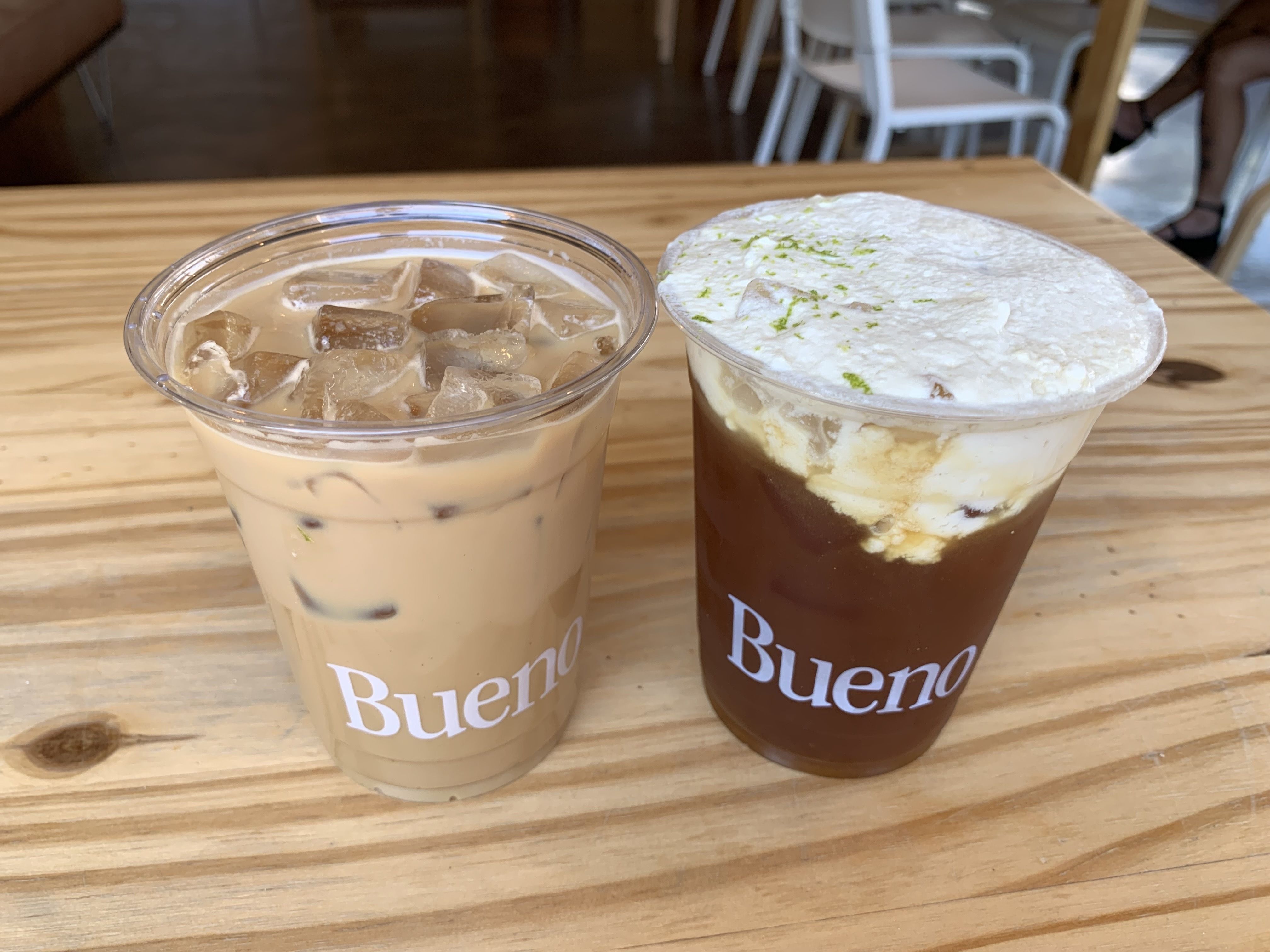Two iced coffee drinks on a wooden table in clear plastic cups labeled "Bueno"; one is light brown with ice and the other is dark with white foam topped with green zest.