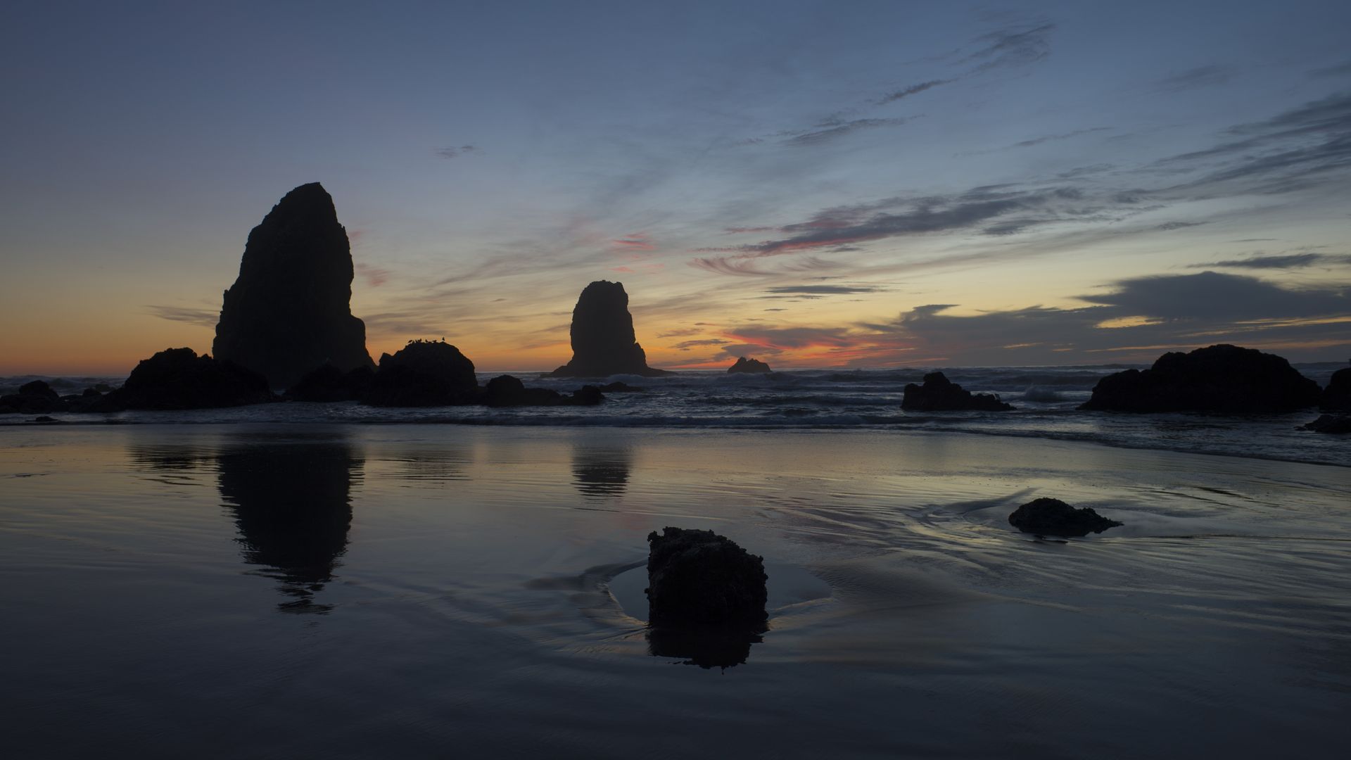 Silhouetted sea stacks rise from calm water at sunset; a gradient sky shifts from deep blue to warm orange near the horizon, with reflections shimmering on the wet sand.