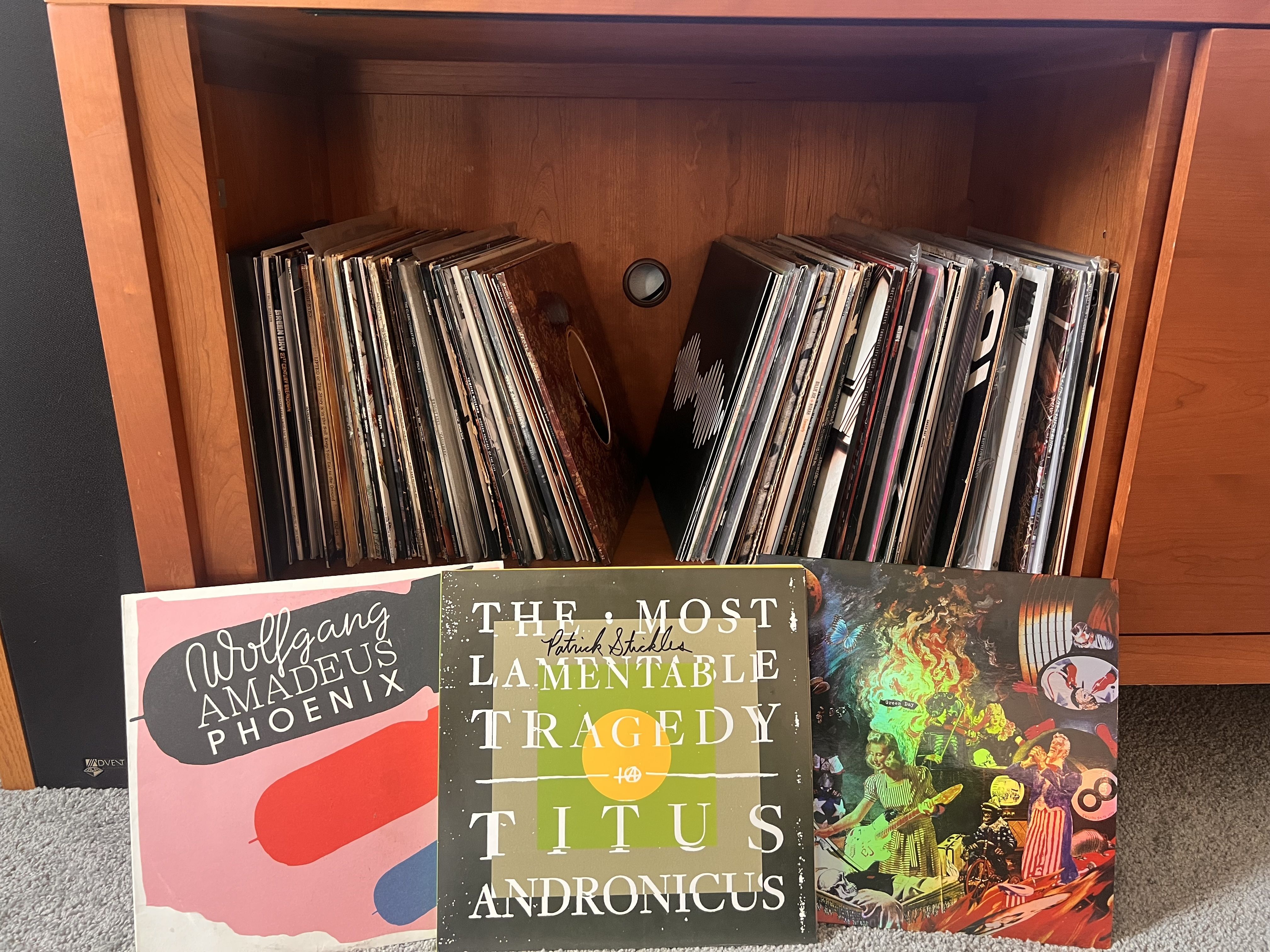 Wooden cabinet filled with vinyl records. Three records are displayed in front: Wolfgang Amadeus Phoenix with pink, red, and black cover; The Most Lamentable Tragedy by Titus Andronicus with text on green background; and a colorful collage album cover.