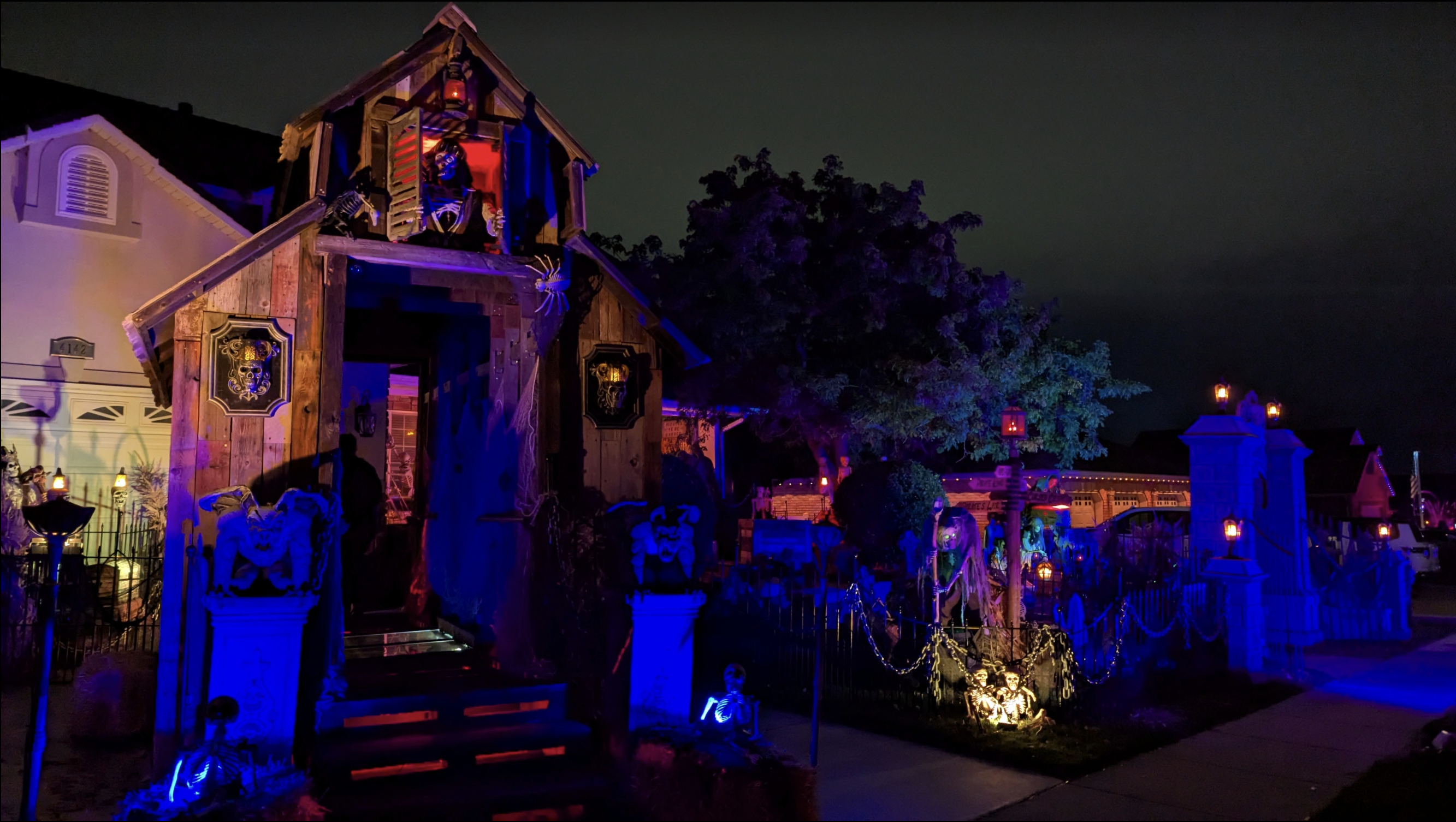Nighttime haunted-house exterior with a wooden façade, skeleton figures visible in the upper window, blue and purple lighting, Halloween decor, and glowing tombstones along a fenced yard.
